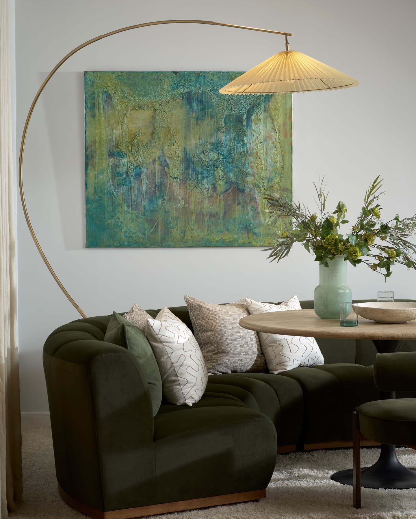 Curved dark green sofa with beige and white cushions, a round table with vases of flowers, a large abstract painting, and an arched standard lamp with a pleated shade.