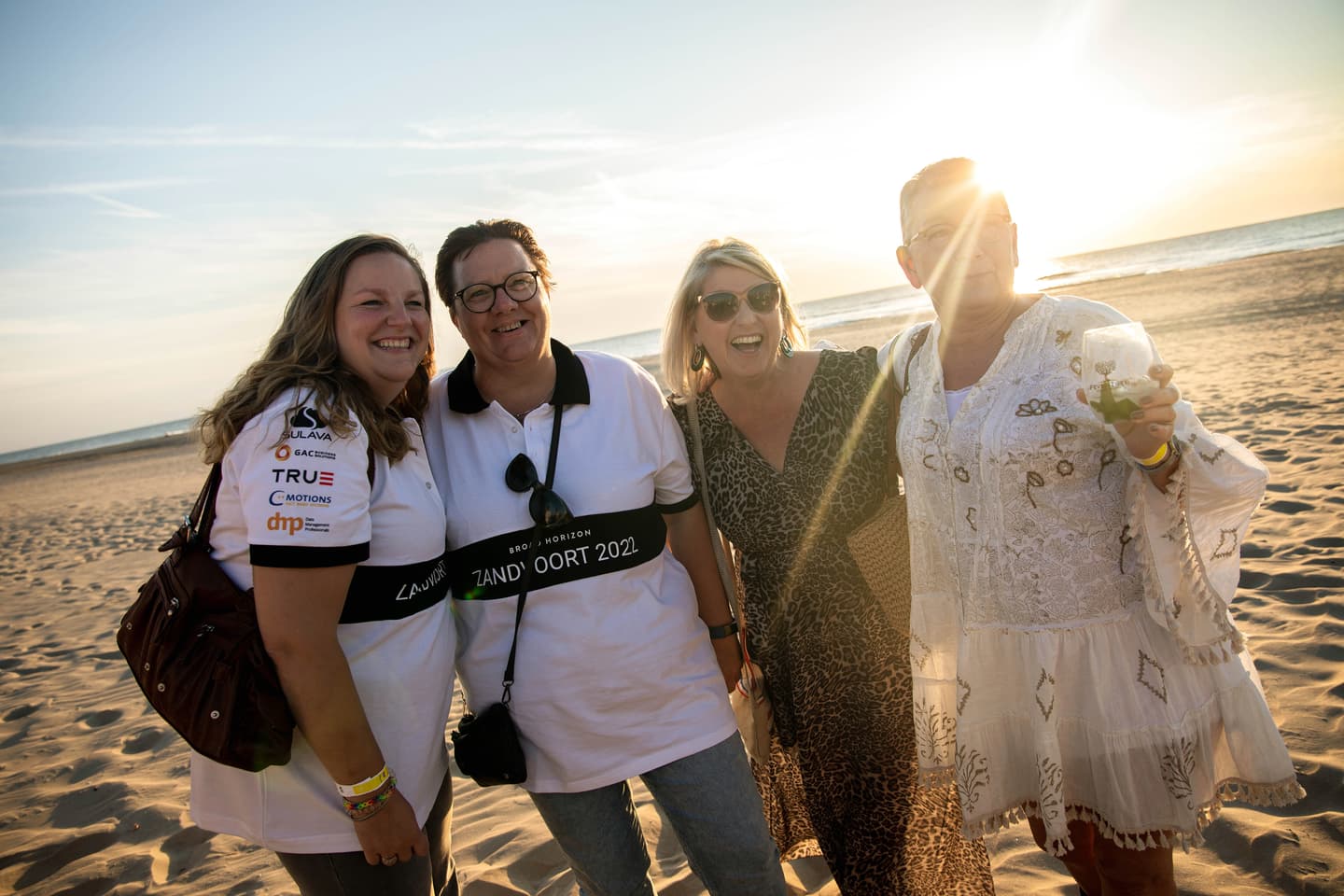 Four women stand smiling together on a sandy beach at sunset, two wearing white shirts, one in a patterned dress, and one in a white lace dress holding a drink.