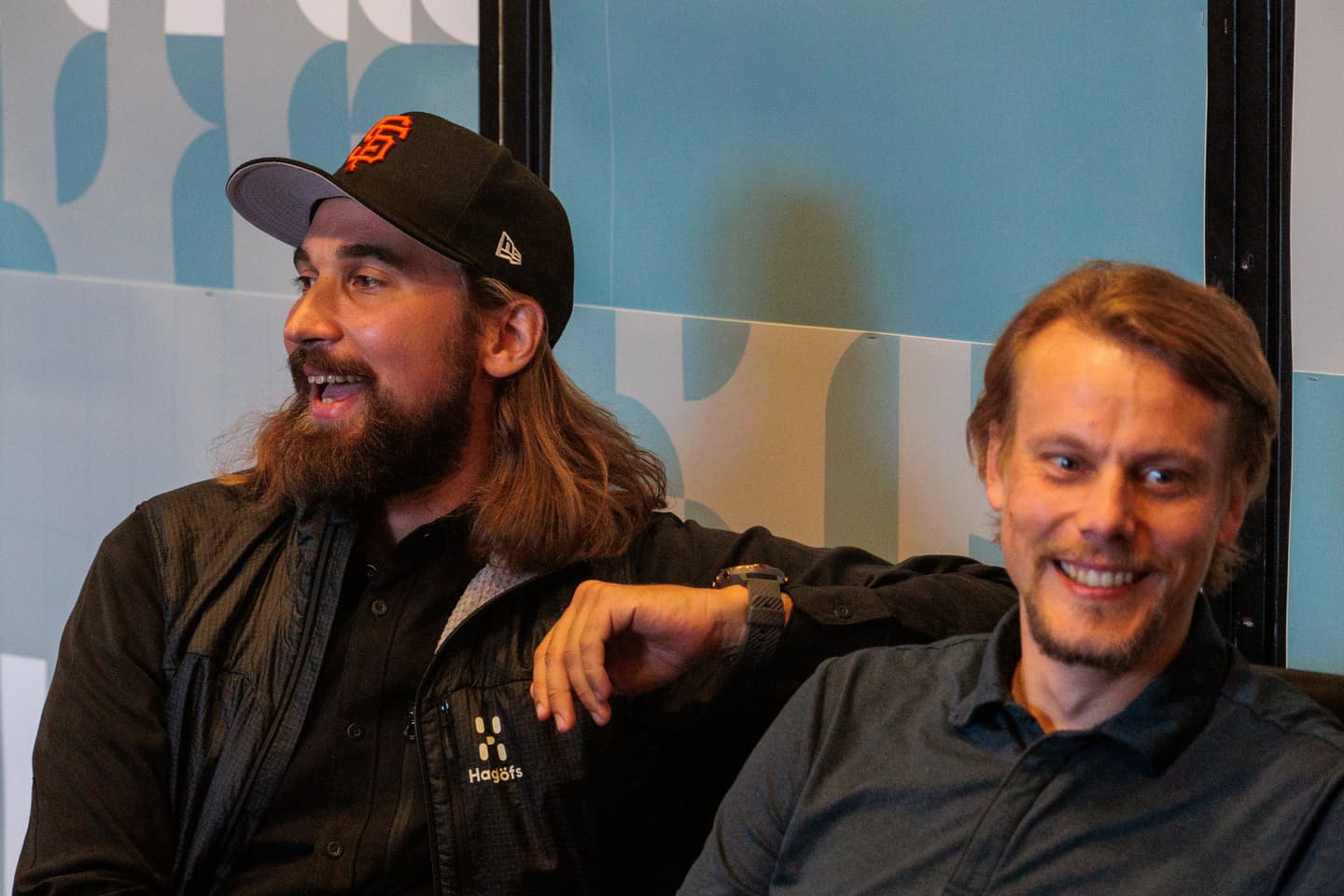 Two men sitting side by side, one wearing a black jacket and baseball cap, both smiling in a brightly lit indoor setting.