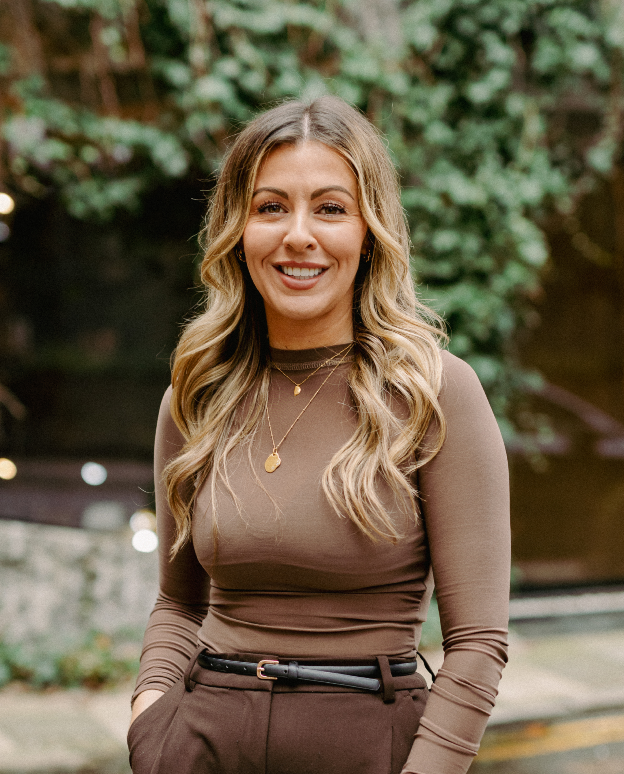 Woman with long wavy hair, wearing a brown long-sleeved top and brown trousers, stands outdoors and smiles at the camera with greenery in the background.