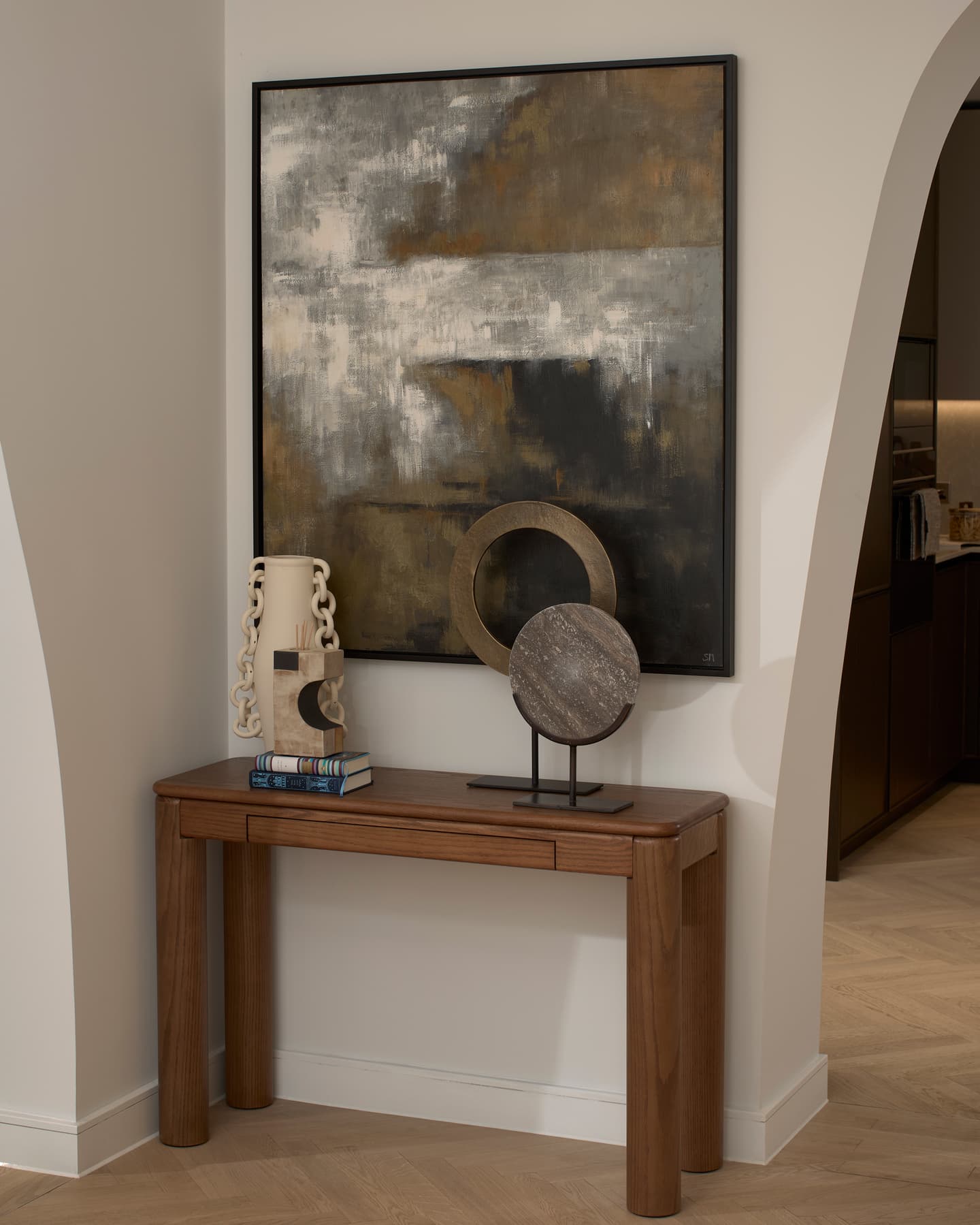 A wooden console table with books, a ceramic vase, and abstract sculptures stands beneath a large abstract painting in a modern corridor.