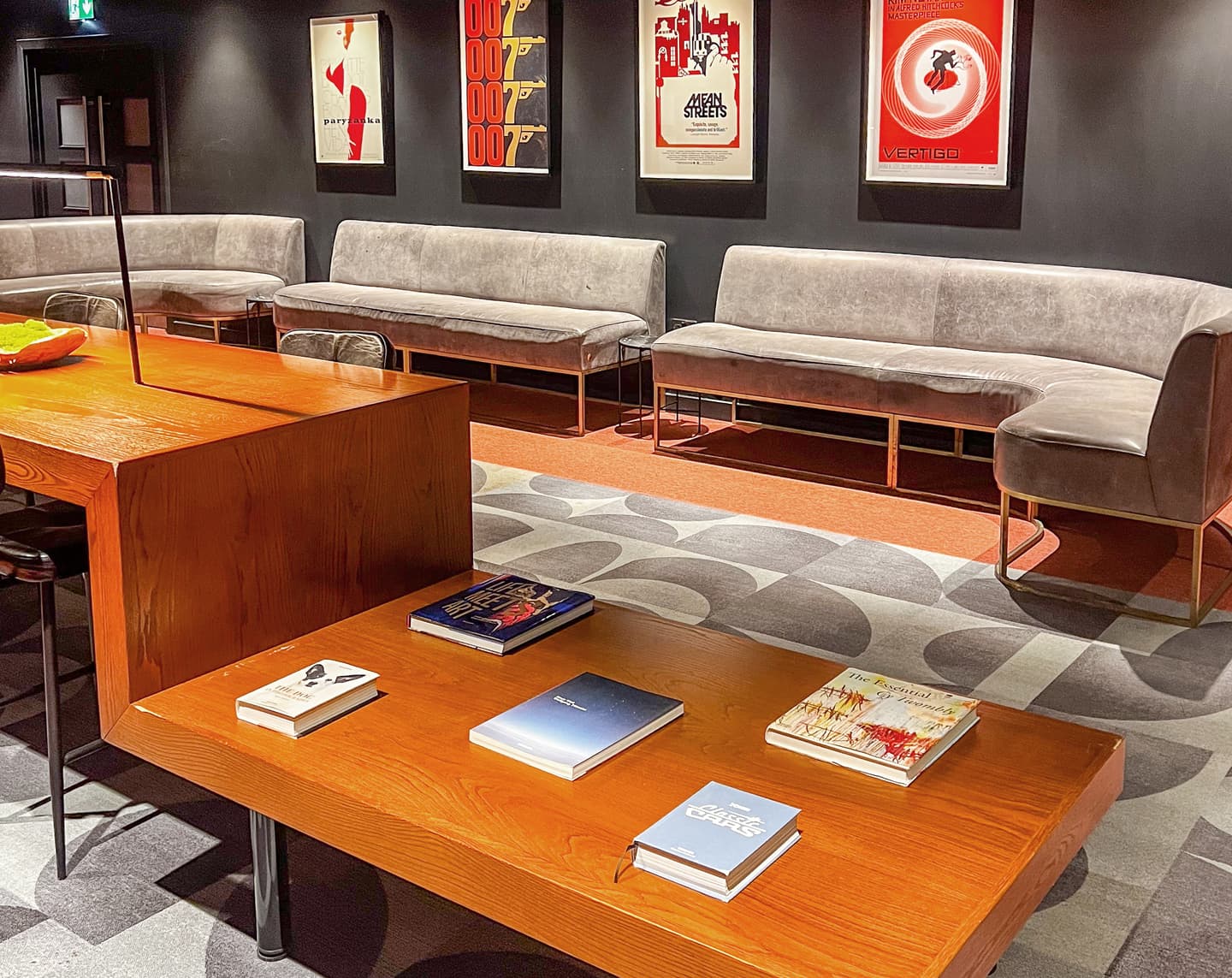 A modern lounge with grey sofas, a wooden coffee table holding books, a patterned rug, and framed vintage posters on a dark wall.