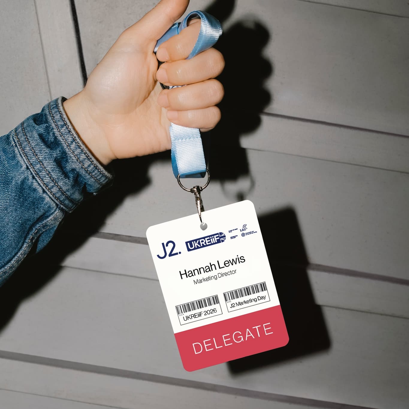 A hand holds a convention badge on a lanyard labelled Hannah Lewis, Marketing Director, DELEGATE, with event details and barcodes.