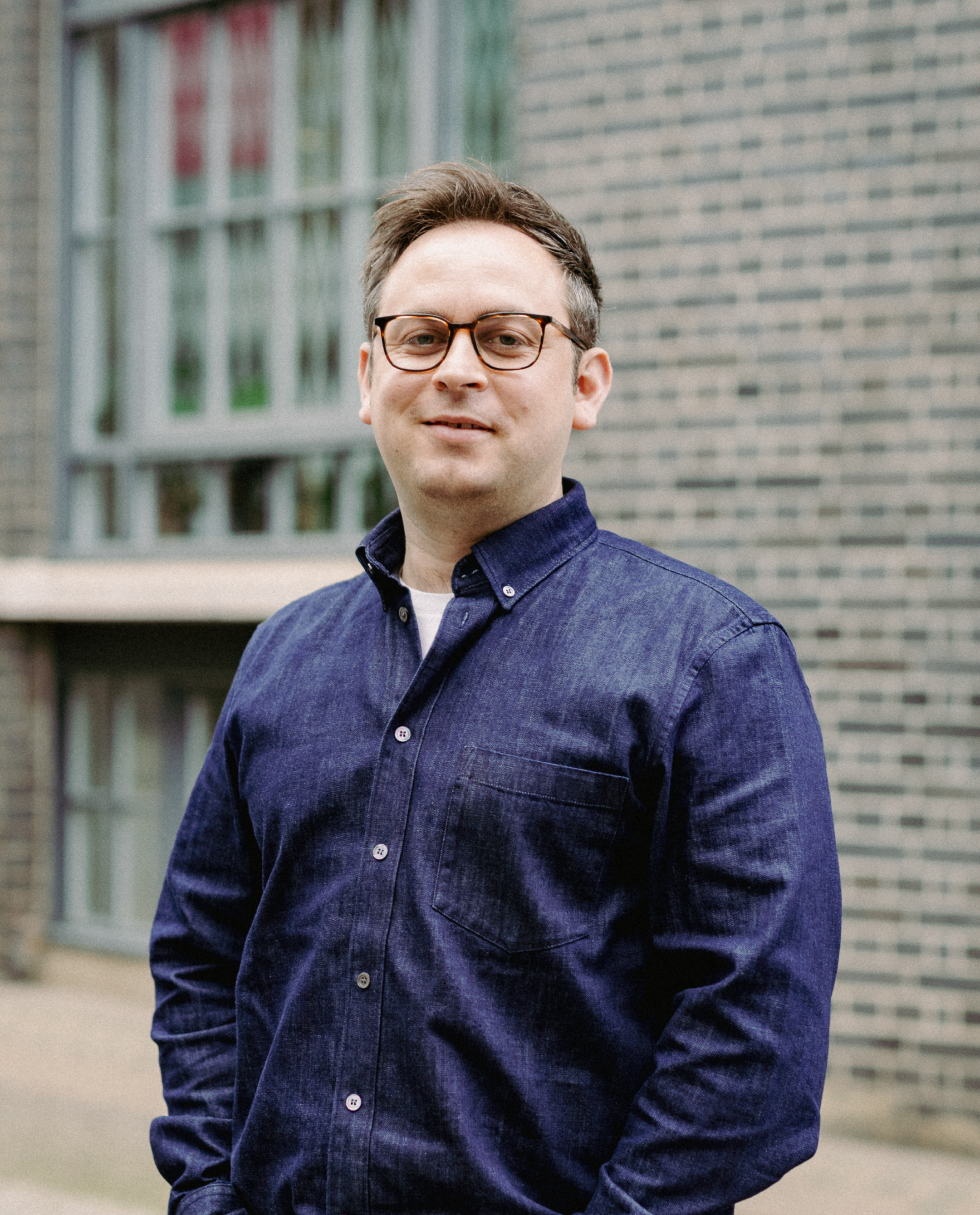 A man wearing glasses and a blue button-up shirt stands outside in front of a grey brick building with large windows.