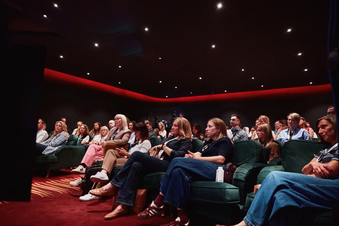 A group of people sits in a dimly lit theatre, attentively watching something off camera.