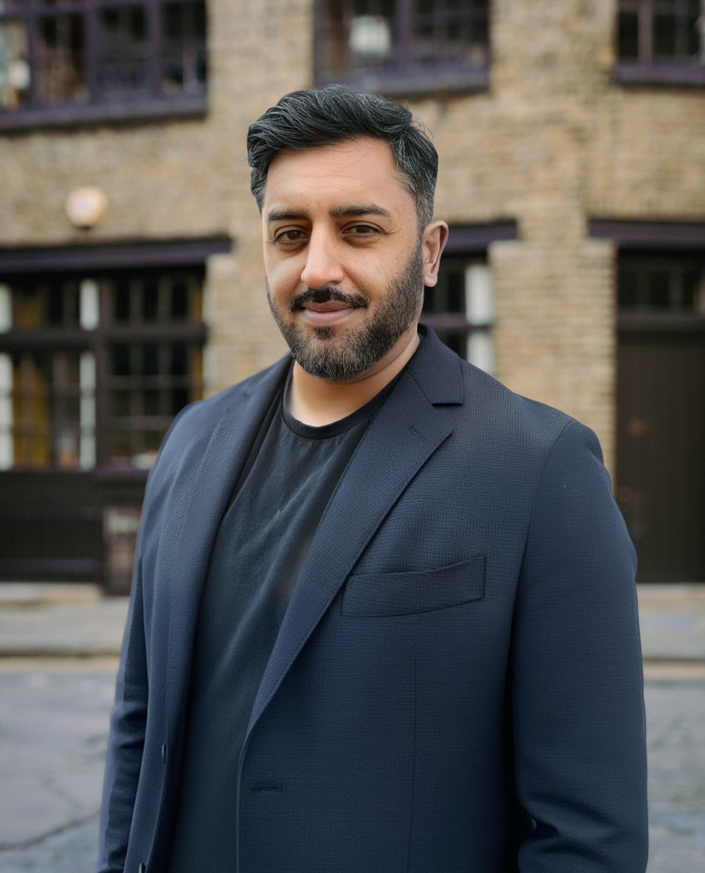 A man with dark hair and a beard wearing a navy blazer and black shirt stands outside in front of a brick building.