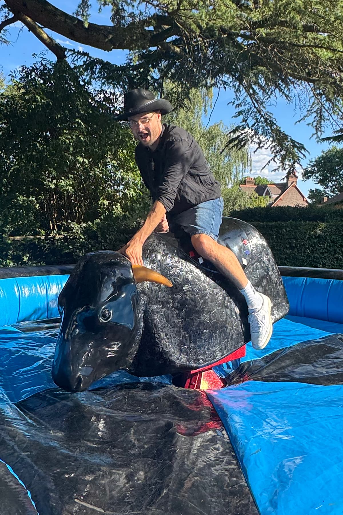 A man wearing a cowboy hat rides a mechanical bull outdoors on a sunny day, surrounded by trees and a blue inflatable safety mat.