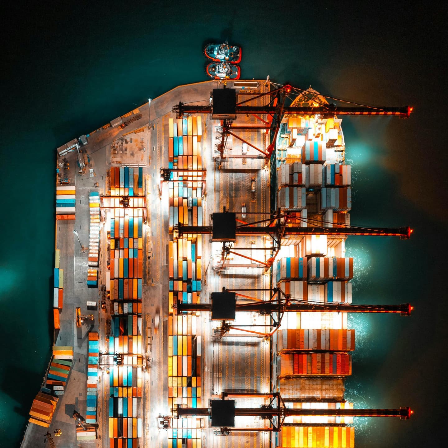 Aerial view of a brightly lit cargo port at night, with shipping containers arranged in rows and large cranes positioned along the quay.