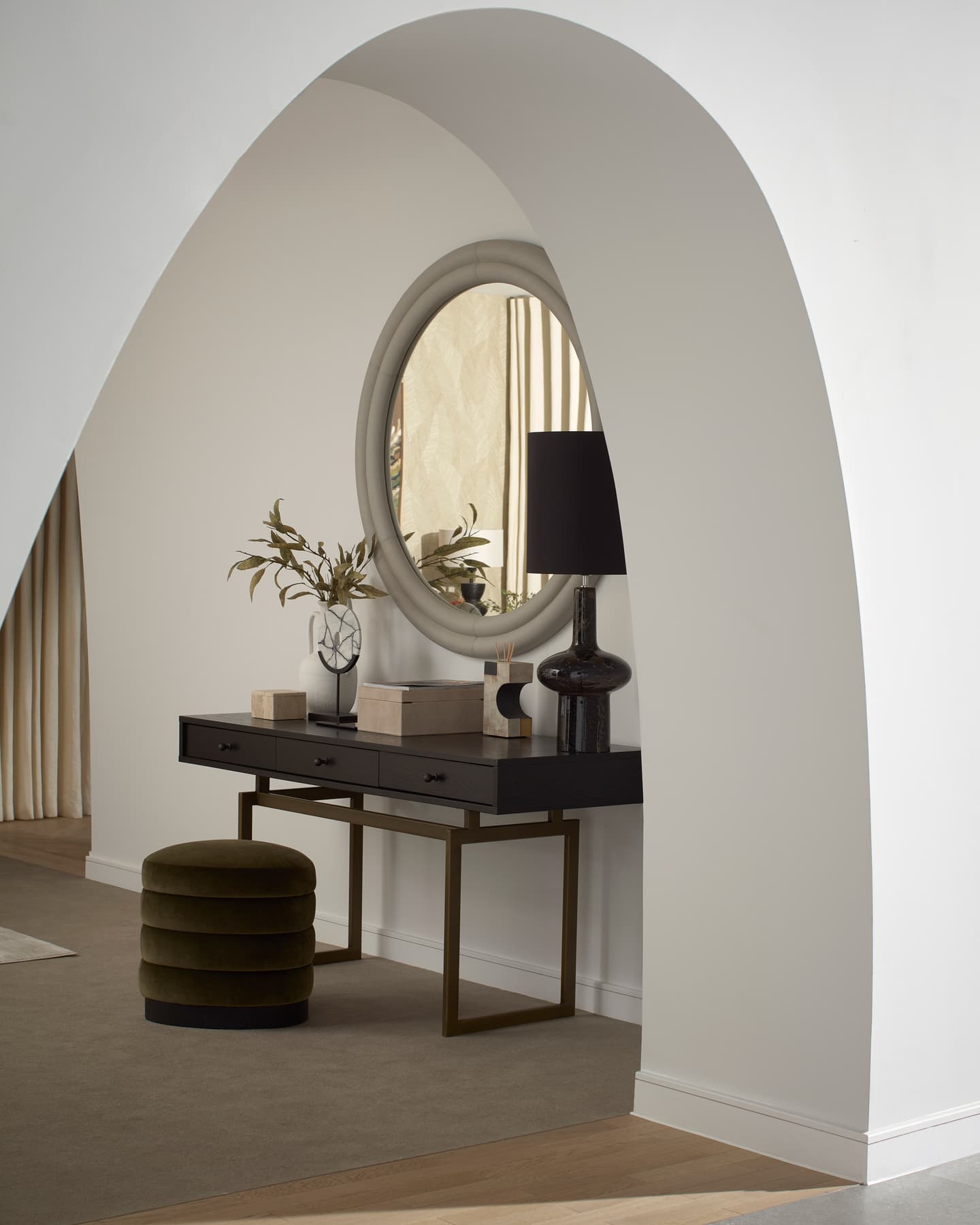 A black console table with decor, a round mirror, lamp, and green ottoman sits beneath a white archway in a modern interior space.