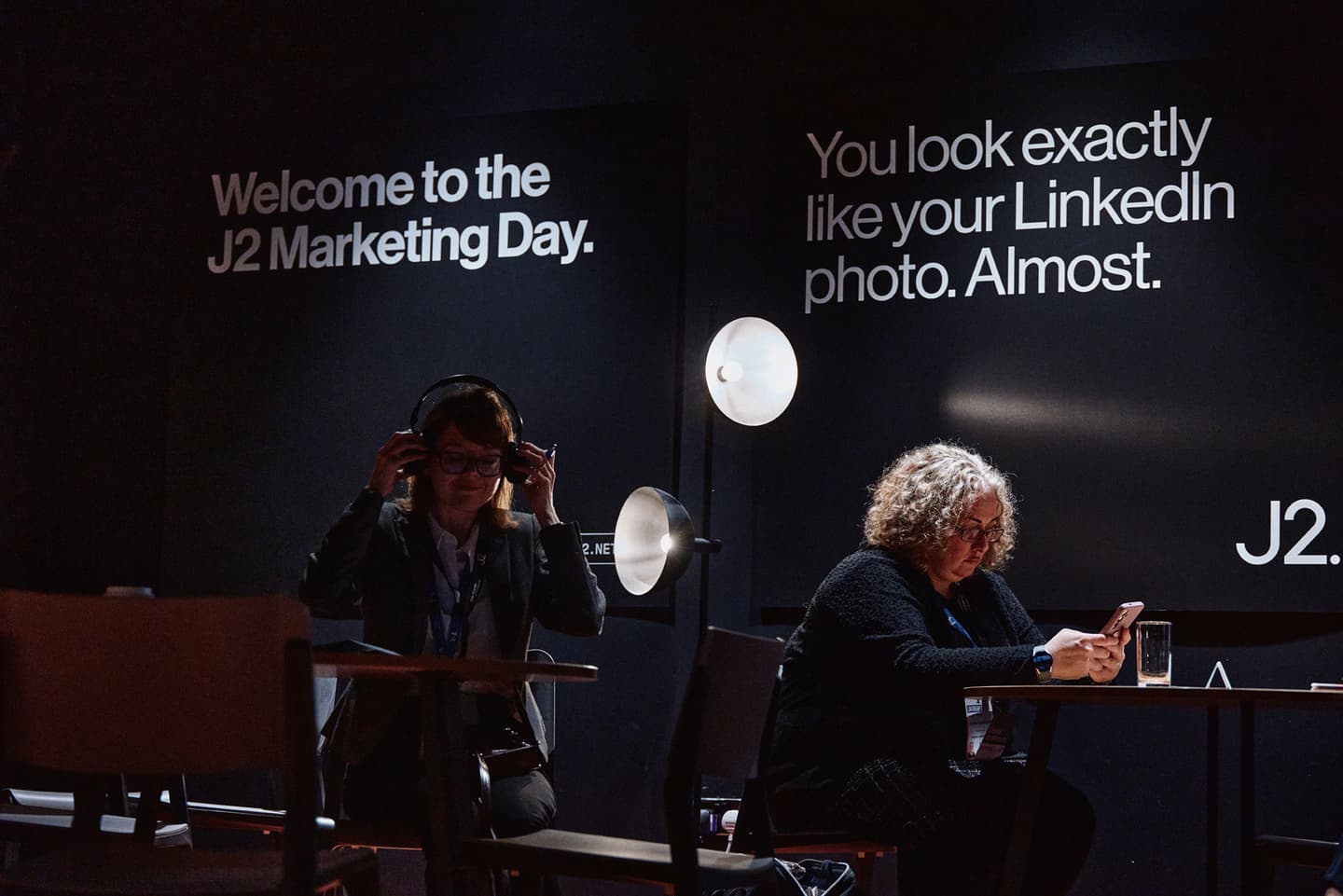 Two people sit at separate tables in a dimly lit room with black walls displaying marketing event signs; one uses a mobile, the other adjusts headphones.
