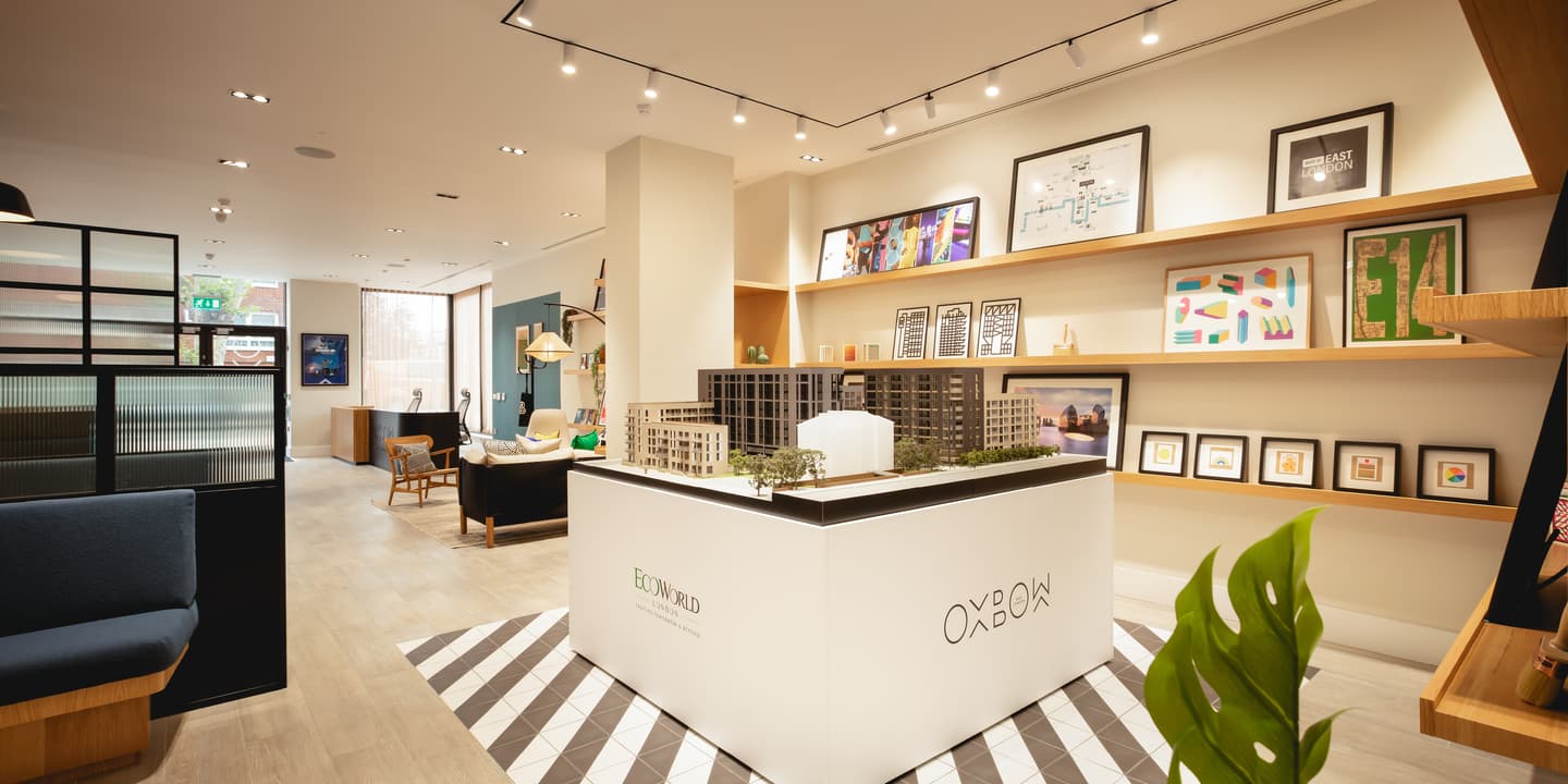 Modern interior showroom with a central architectural model display, framed artwork on shelves, contemporary furniture, and bright lighting.