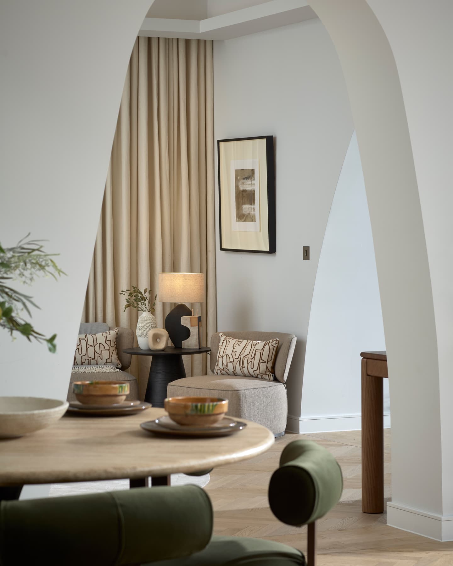 A modern living space with neutral colours, arched doorways, a round table with green chairs, a lamp, and abstract wall art beside a window with beige curtains.