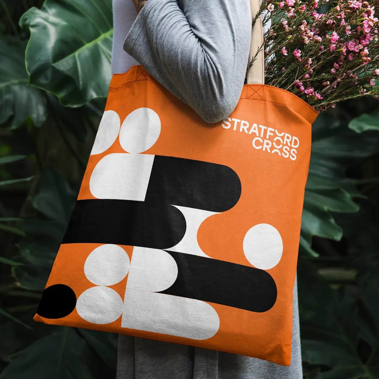 Person holding an orange tote bag with abstract black and white geometric shapes and the text “STRATFORD CROSS,” carrying pink flowers.