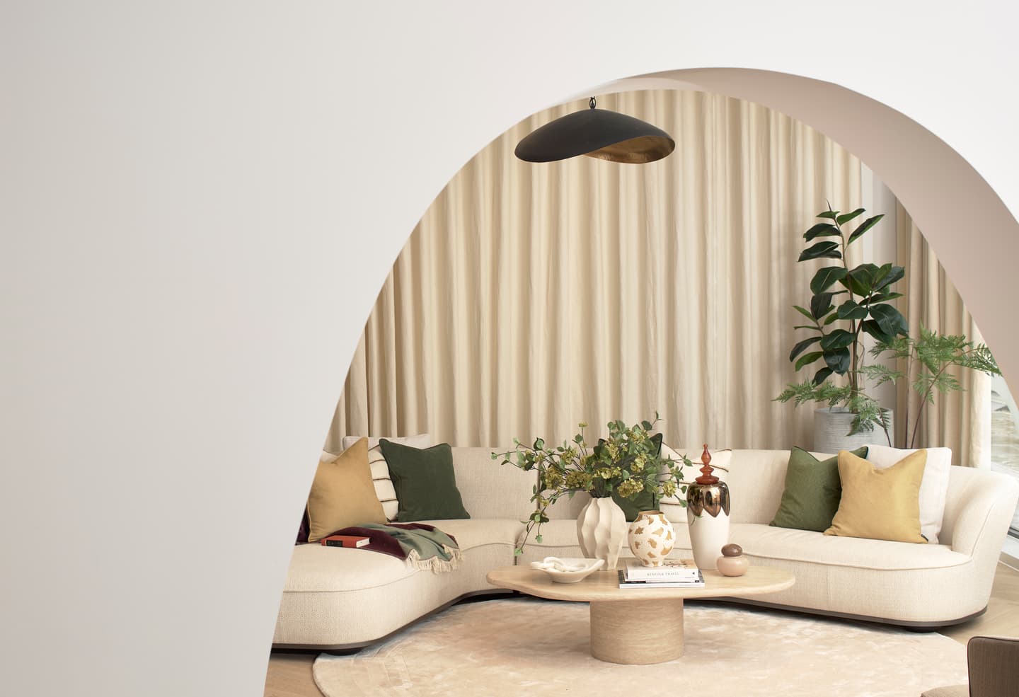 Contemporary living room with a curved light sofa, assorted cushions, a round coffee table with decor, potted plants, and beige curtains, viewed through a white arched opening.