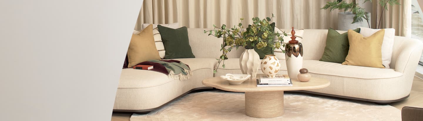 A modern living room with a light beige corner sofa, green and tan cushions, a wooden coffee table with decor items, a beige rug, and neutral curtains in the background.