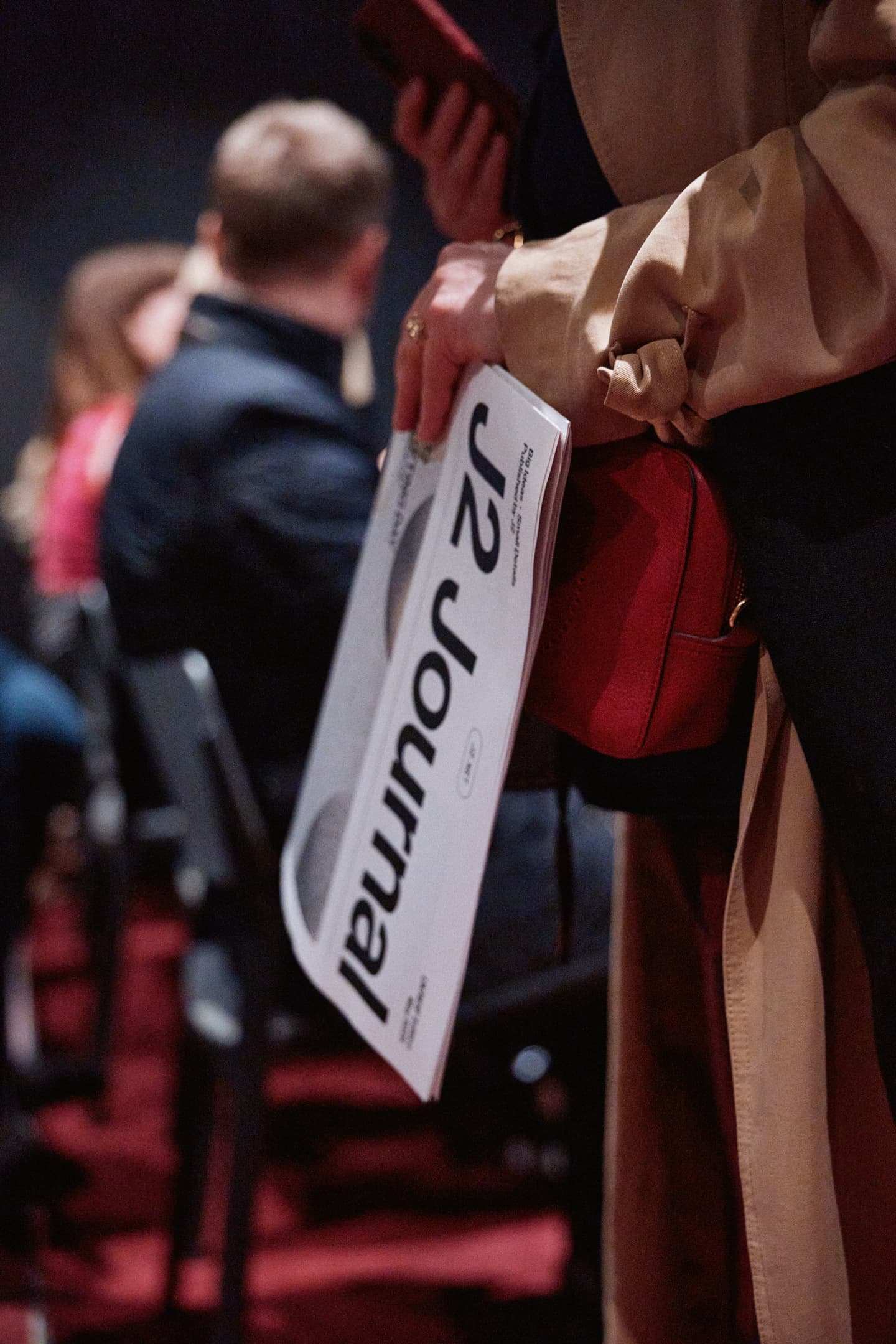 Person holding a folded J2 Journal in one hand and a red bag, with others seated in the background.