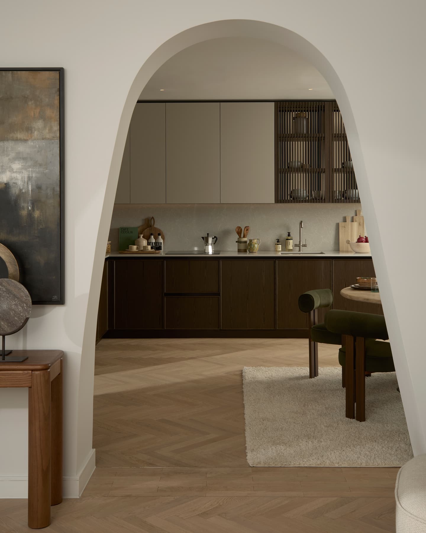 A modern kitchen with dark lower cupboards, light upper cupboards, and minimalist decor is viewed through an archway from a dining area with a wooden table and beige rug.