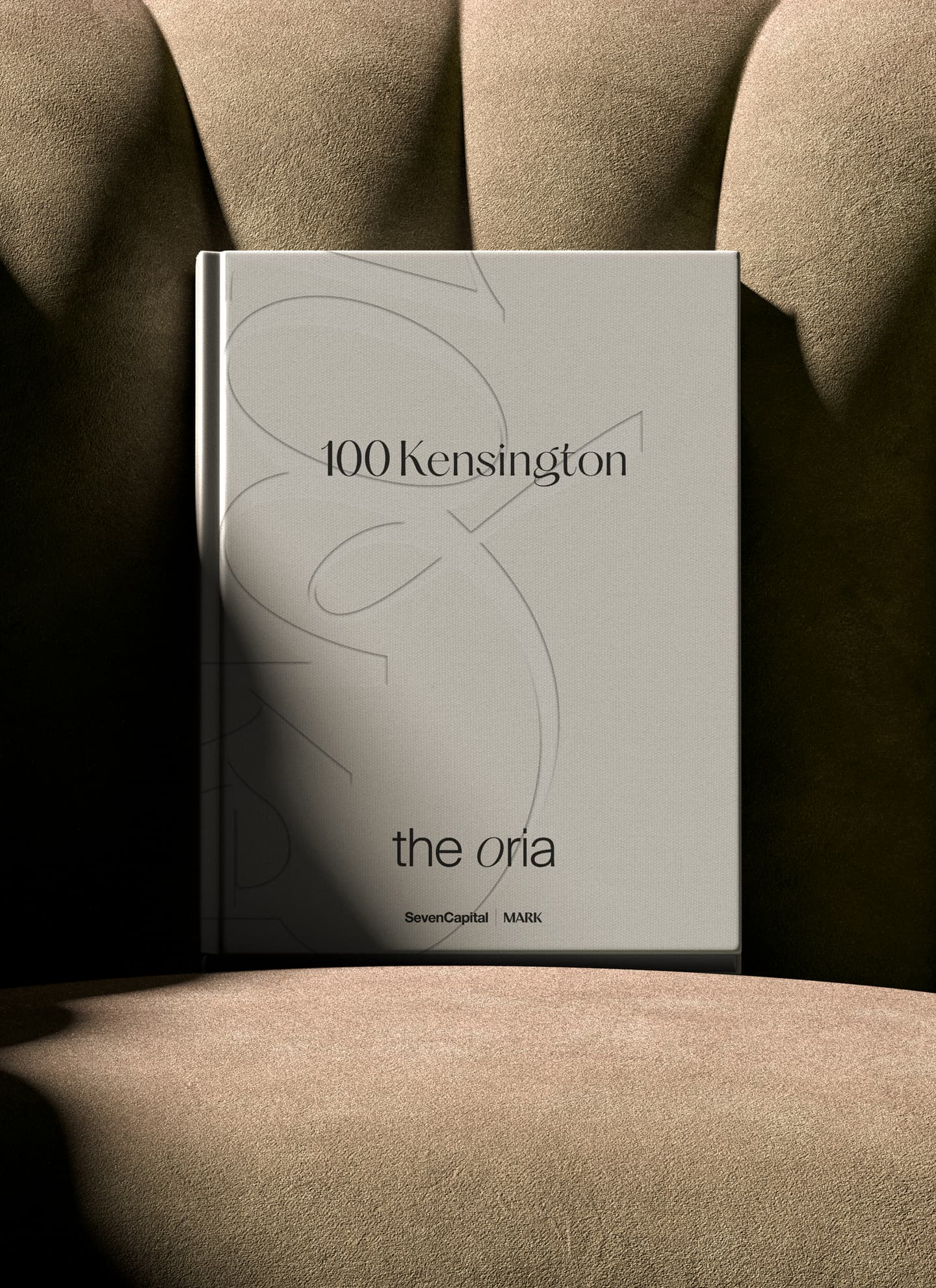 A hardback book titled 100 Kensington the oria rests upright on a beige upholstered chair; the cover also features the logos of SevenCapital and MARK.