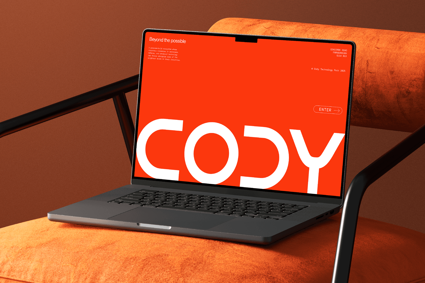 A laptop on an orange chair displays a bright red screen with large white text reading CODY and a small ENTER button on the right-hand side.