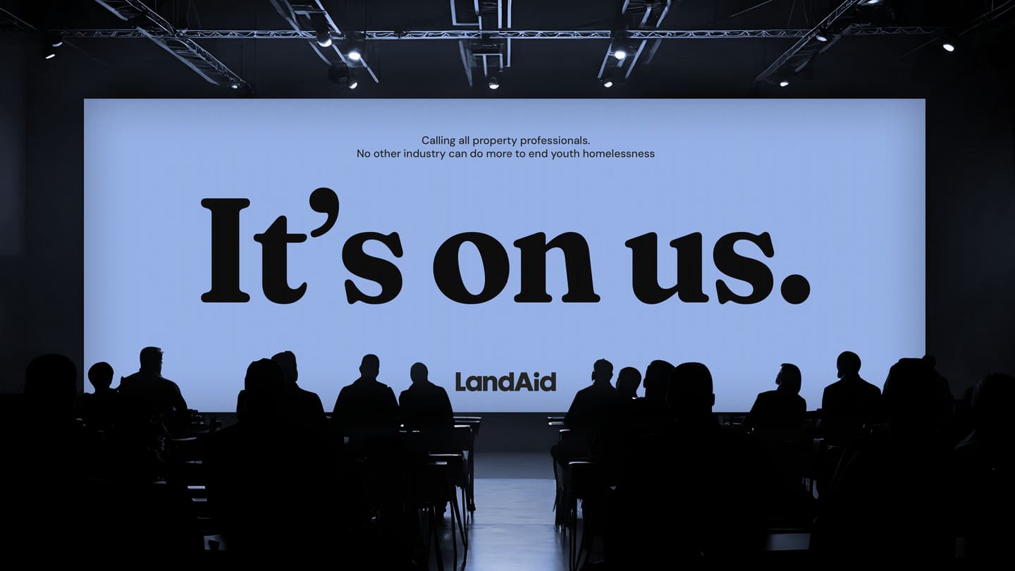Silhouetted audience faces a large screen displaying the message It’s on us. with smaller text calling property professionals to address youth homelessness. LandAid logo shown.