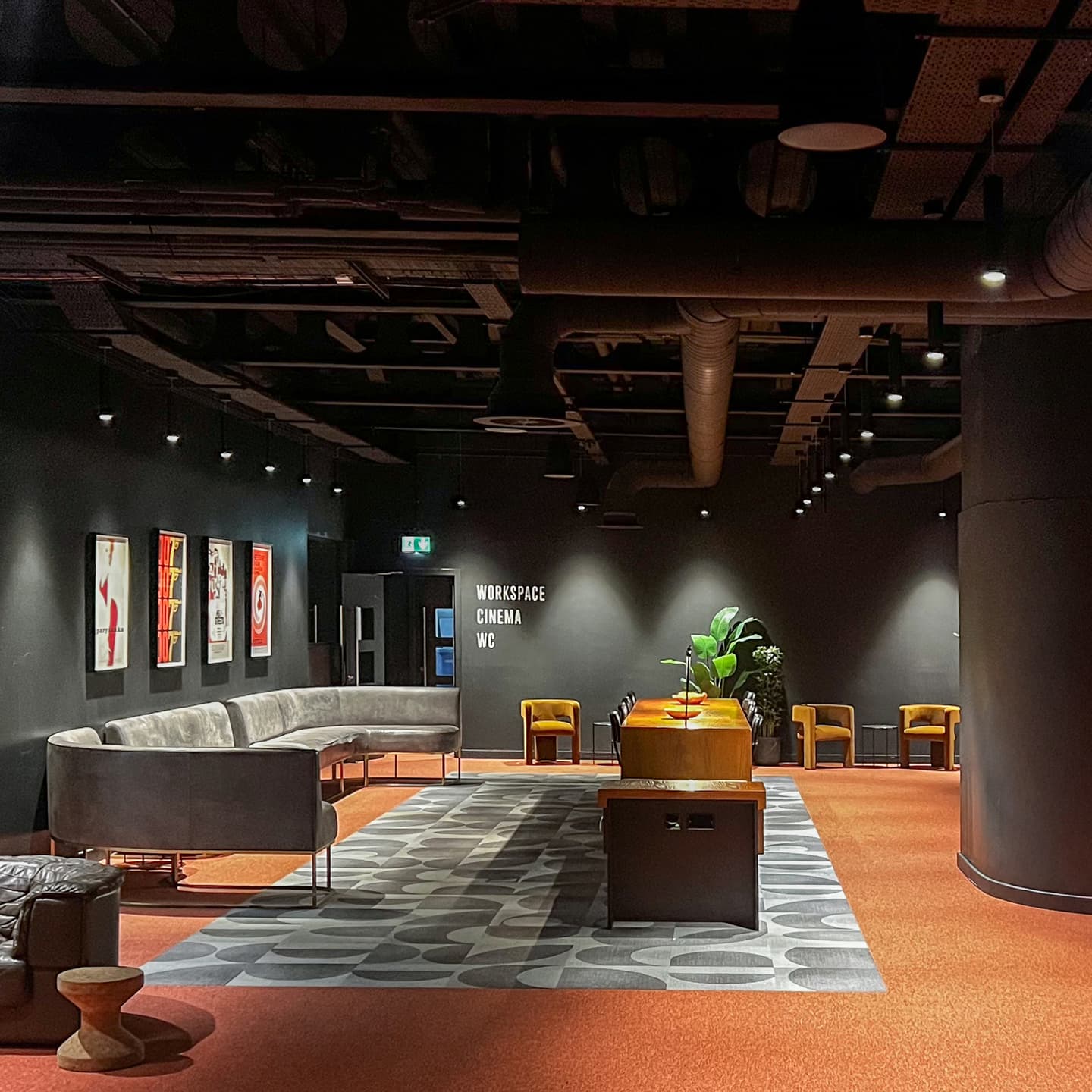 Modern lounge area with grey sofas, small tables, wall art, and a patterned rug, featuring signs for workspace, cinema, and WC on a dark wall.