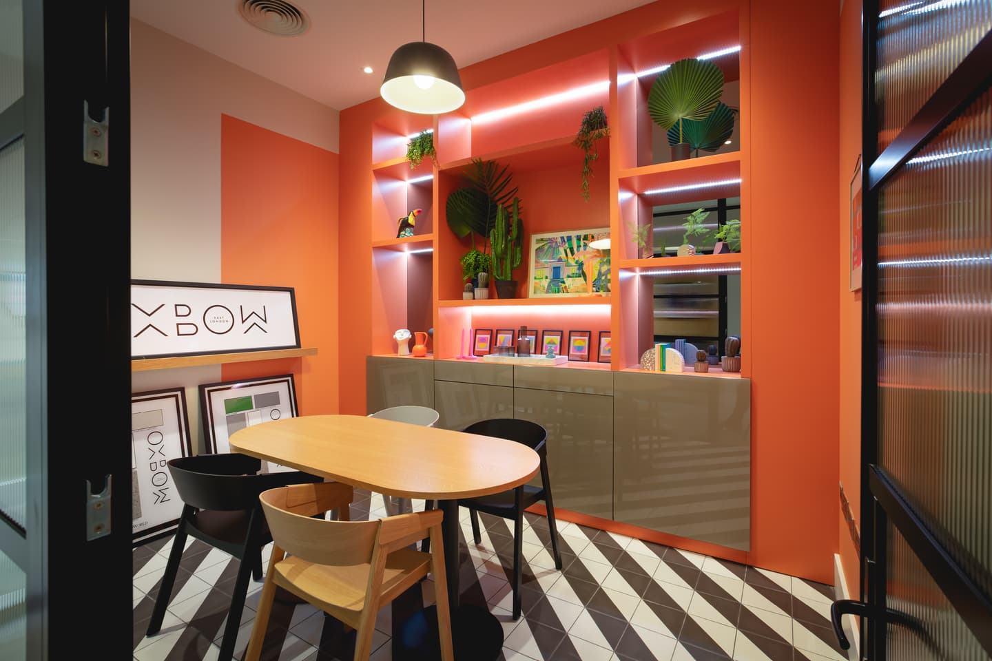 A modern meeting room with an oval table, mixed chairs, geometric flooring, coral walls, and illuminated shelves displaying books, plants, and framed artwork.