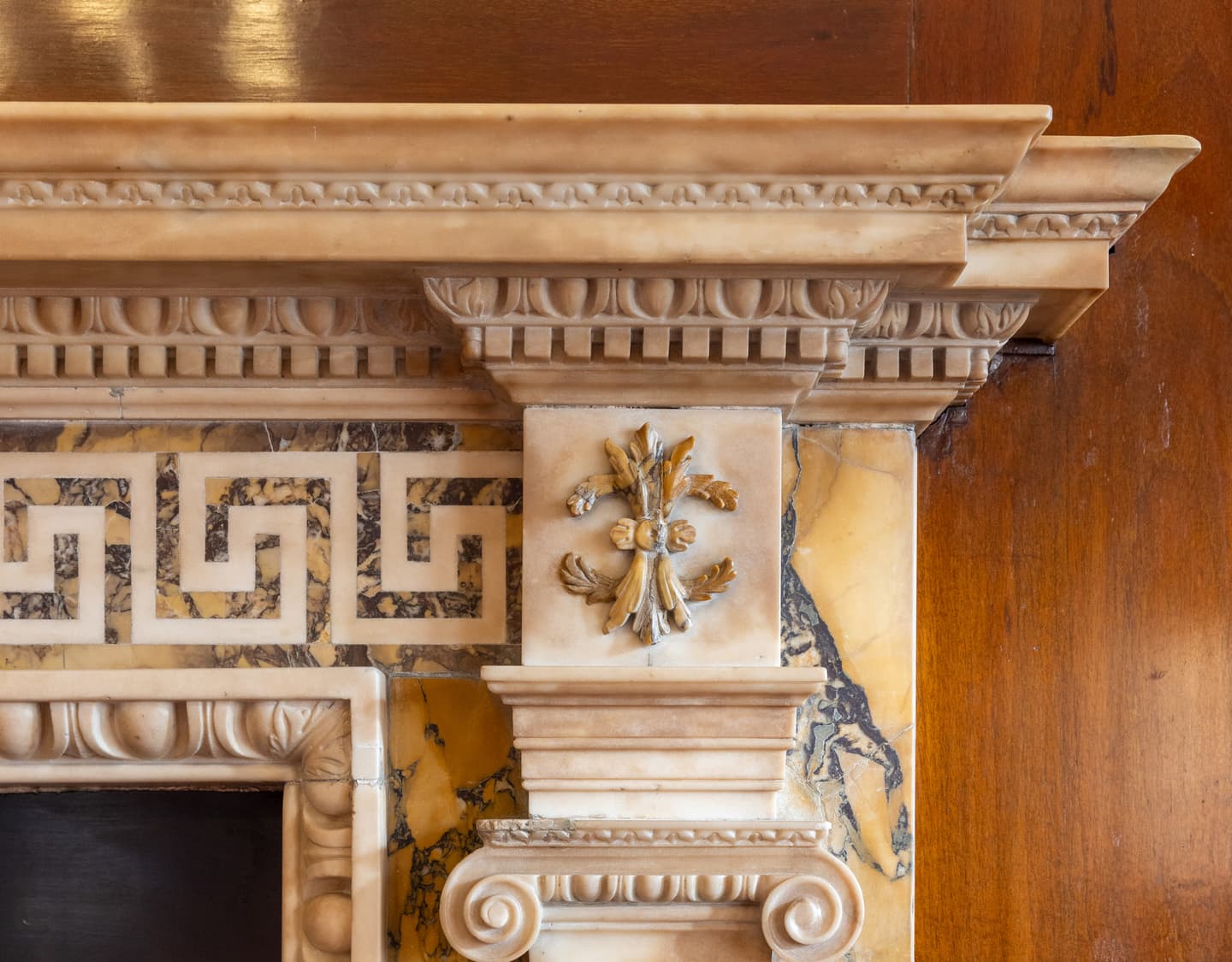 Close-up of an ornate marble fireplace with Greek key pattern, decorative trim, and a gold floral accent on a polished brown and yellow stone background.