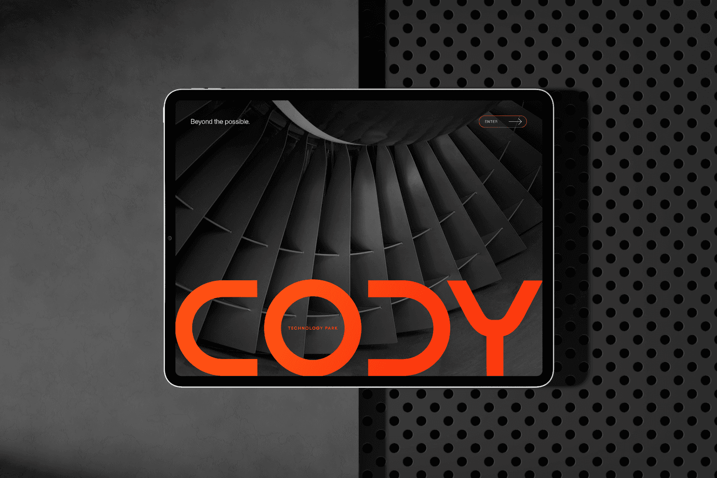 A tablet displays a close-up image of turbine blades with the word CODY in large orange letters across the screen, lying on a two-toned, perforated metal surface.