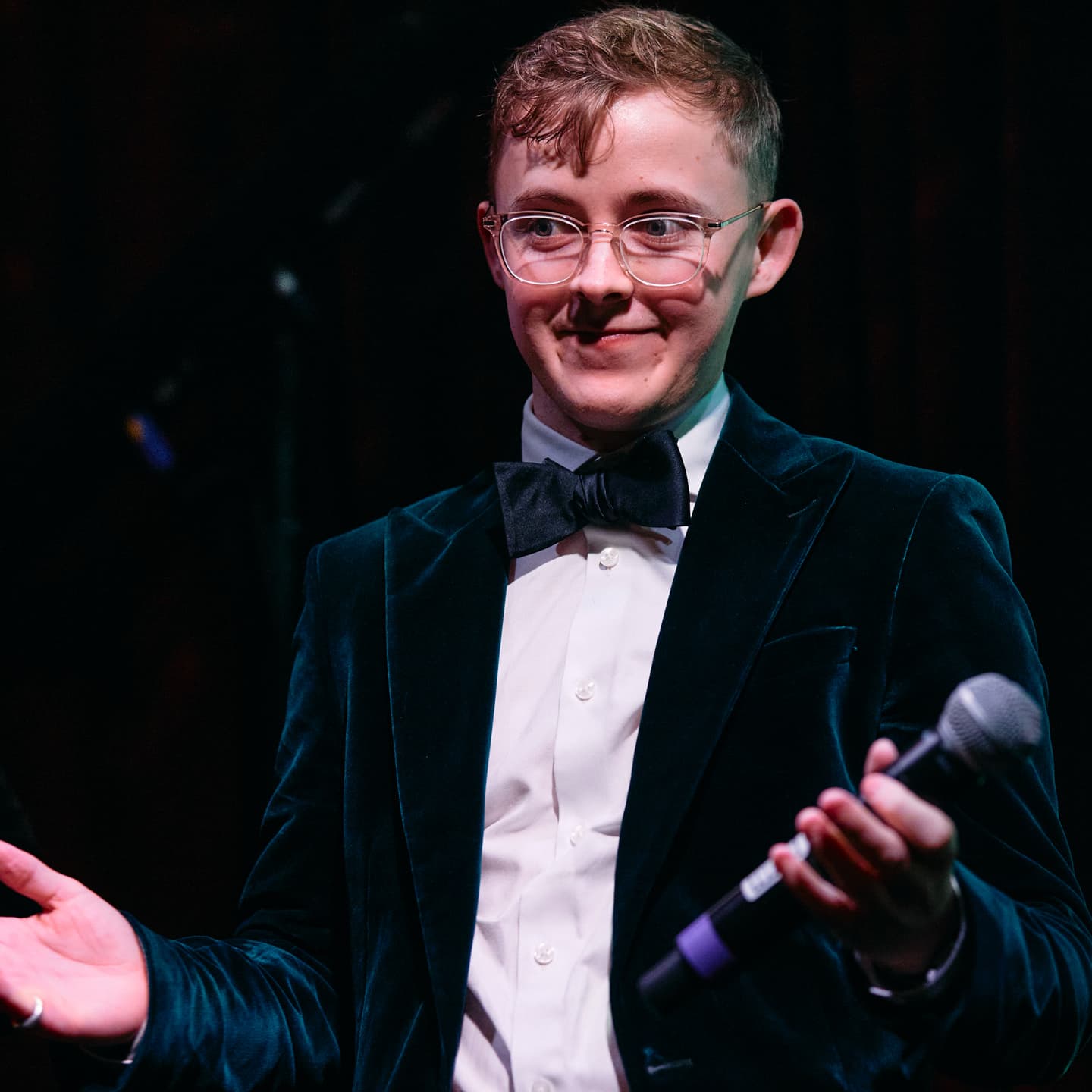 A person in a velvet suit, white shirt, and bow tie holds a microphone and gestures with one hand, standing on stage.