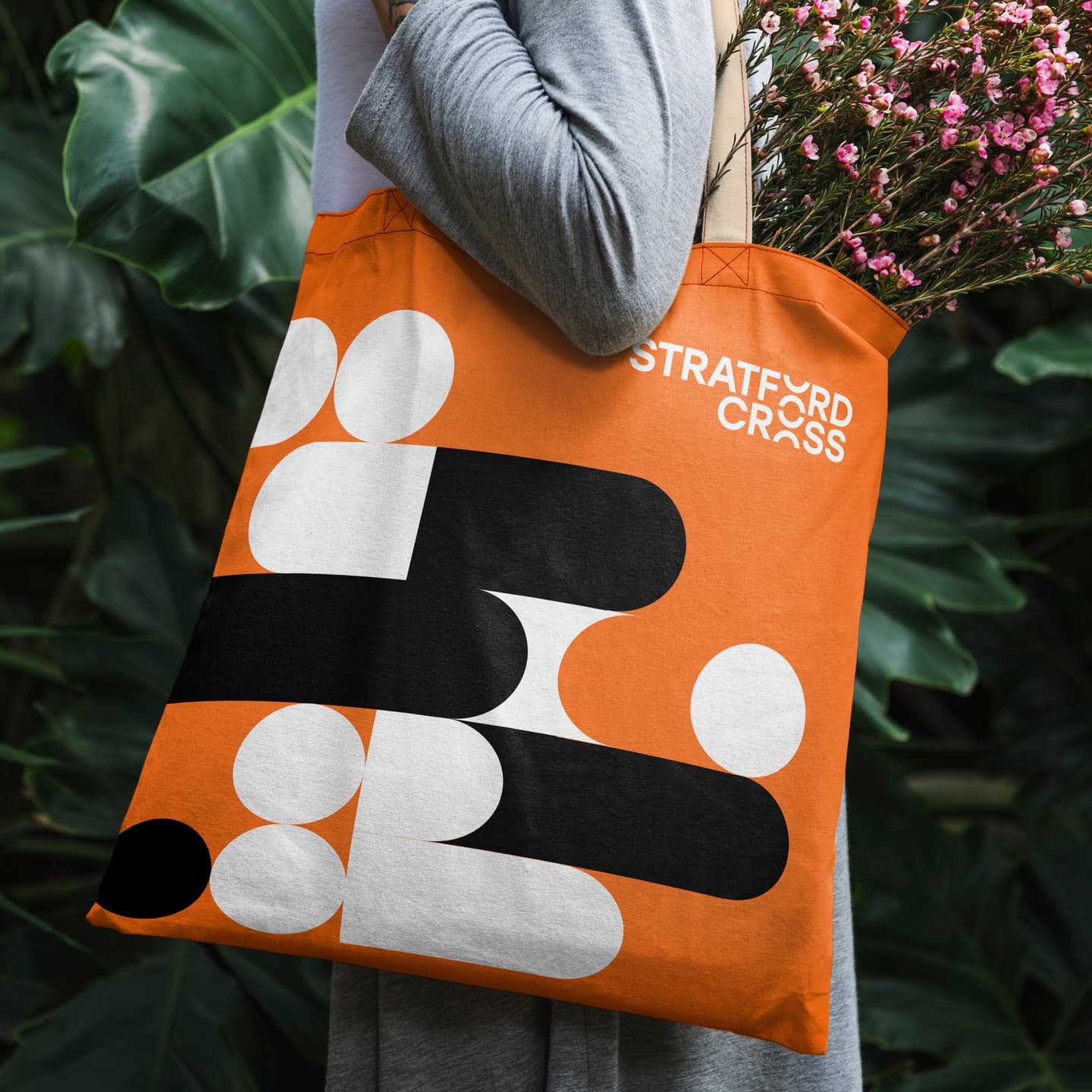 Person holding an orange tote bag with abstract black and white shapes and the text Stratford Cross, containing a bunch of pink flowers. Large green leaves are in the background.