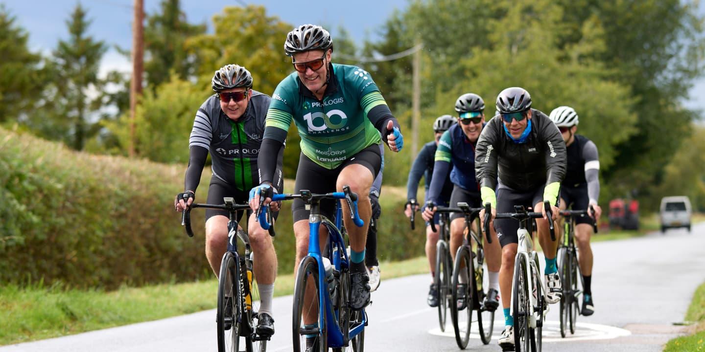A group of cyclists ride together on a rural road, led by a man in a green jersey who is smiling and giving a thumbs-up.