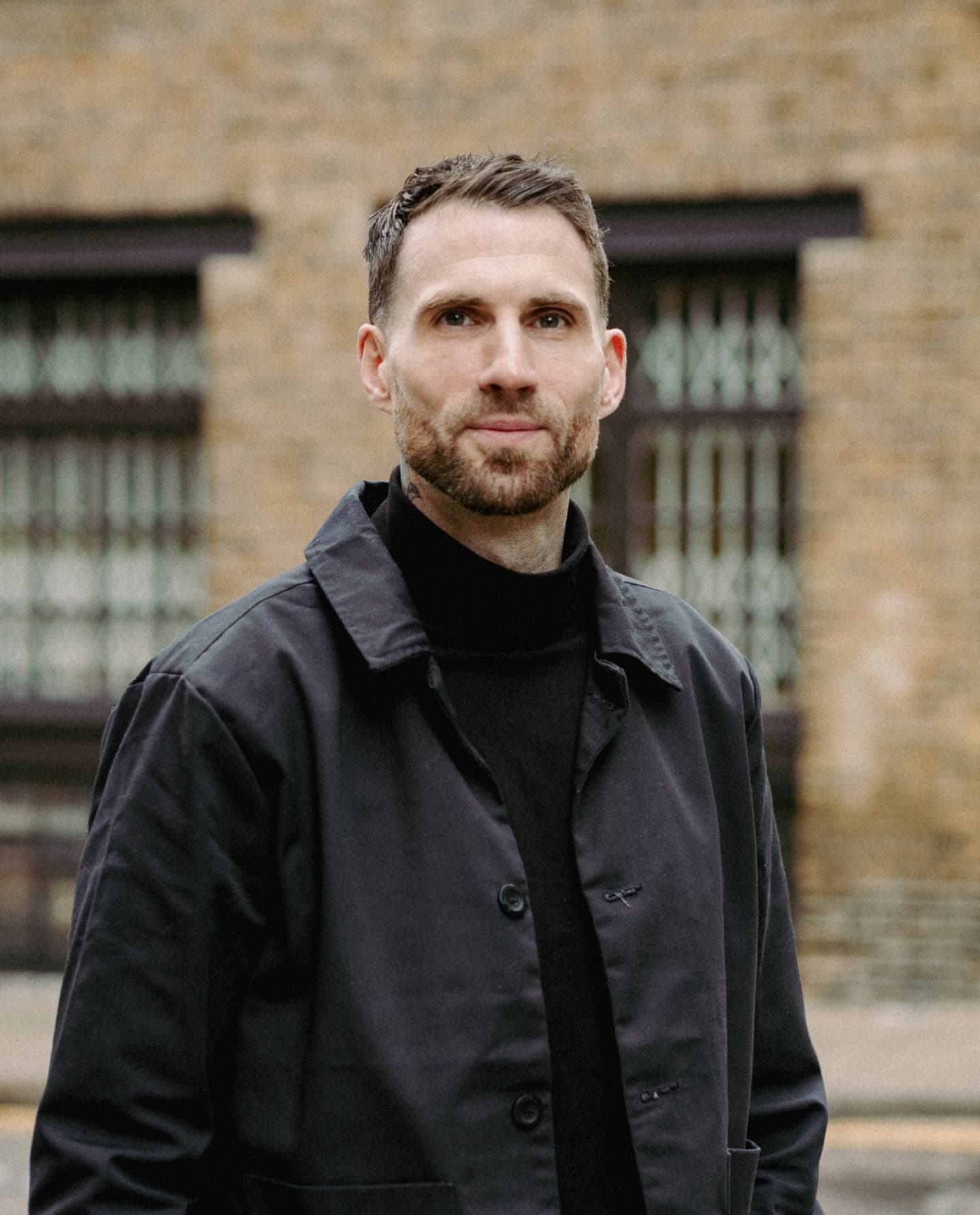 A man with short brown hair and a trimmed beard stands outdoors in front of a brick building, wearing a black coat and polo neck.