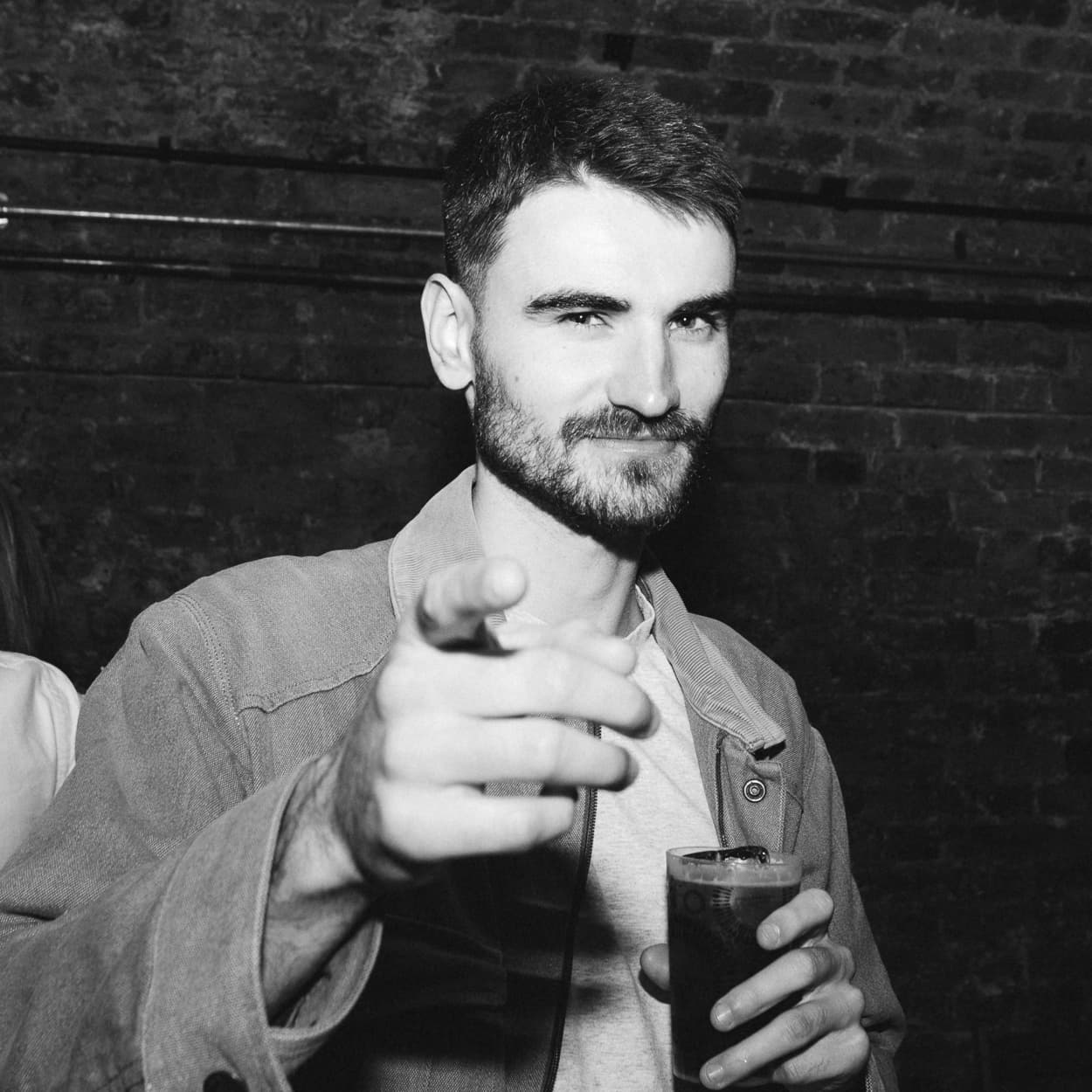 A man with a beard points towards the camera whilst holding a drink, standing in front of a brick wall.