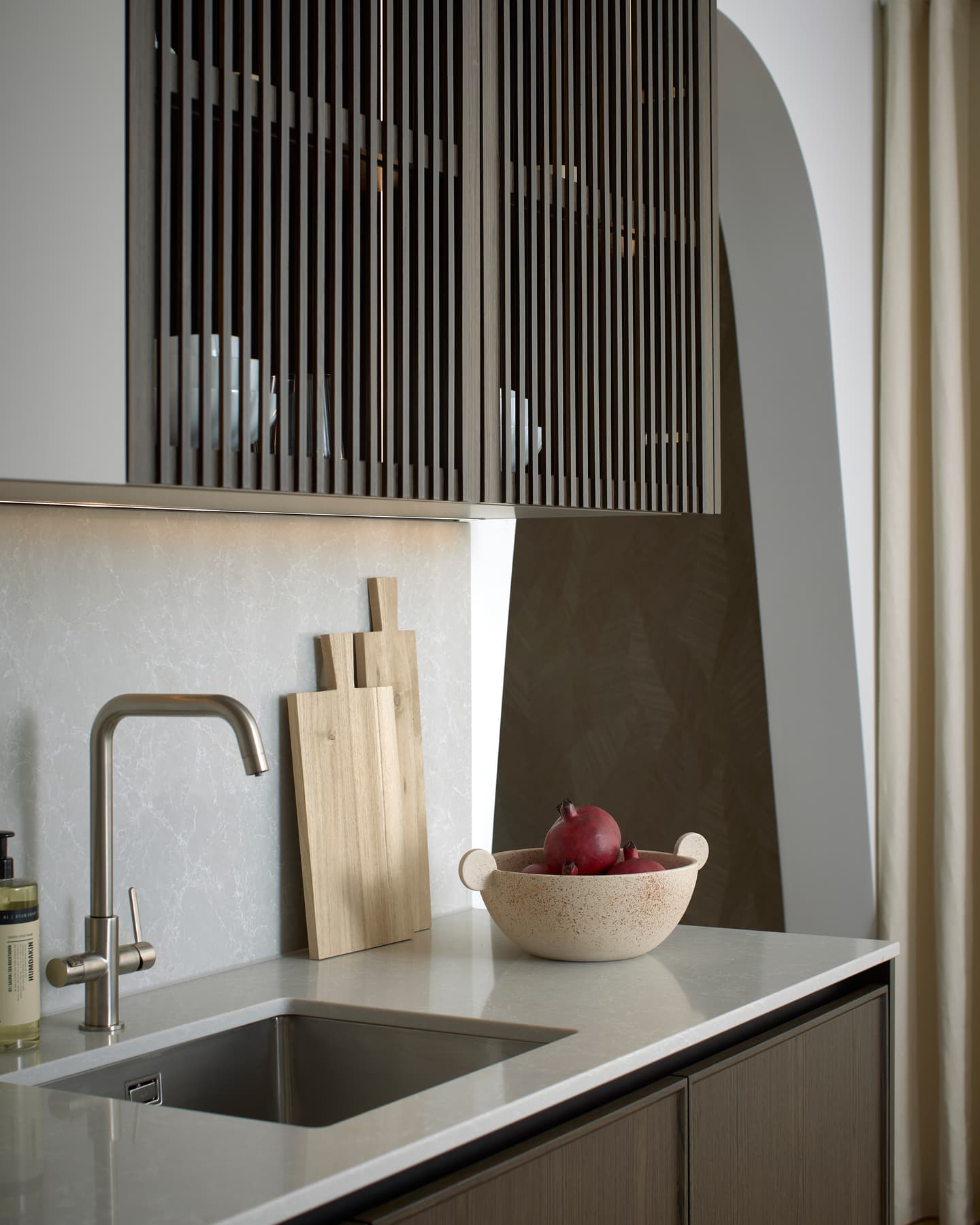 Modern kitchen worktop with a stainless steel sink, tap, two wooden chopping boards, and a bowl containing two pomegranates. Upper cupboards have vertical slatted doors.