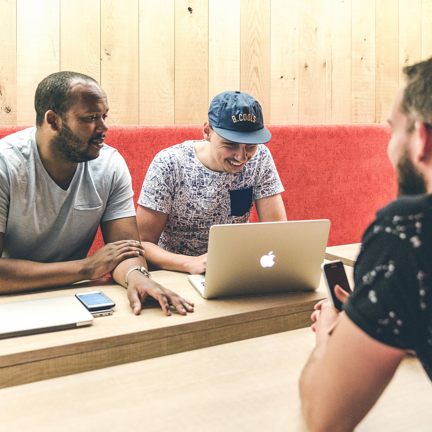 Three men sit at a wooden table, talking and looking at a laptop; two mobile phones are on the table, and the background features a red bench and wooden wall.