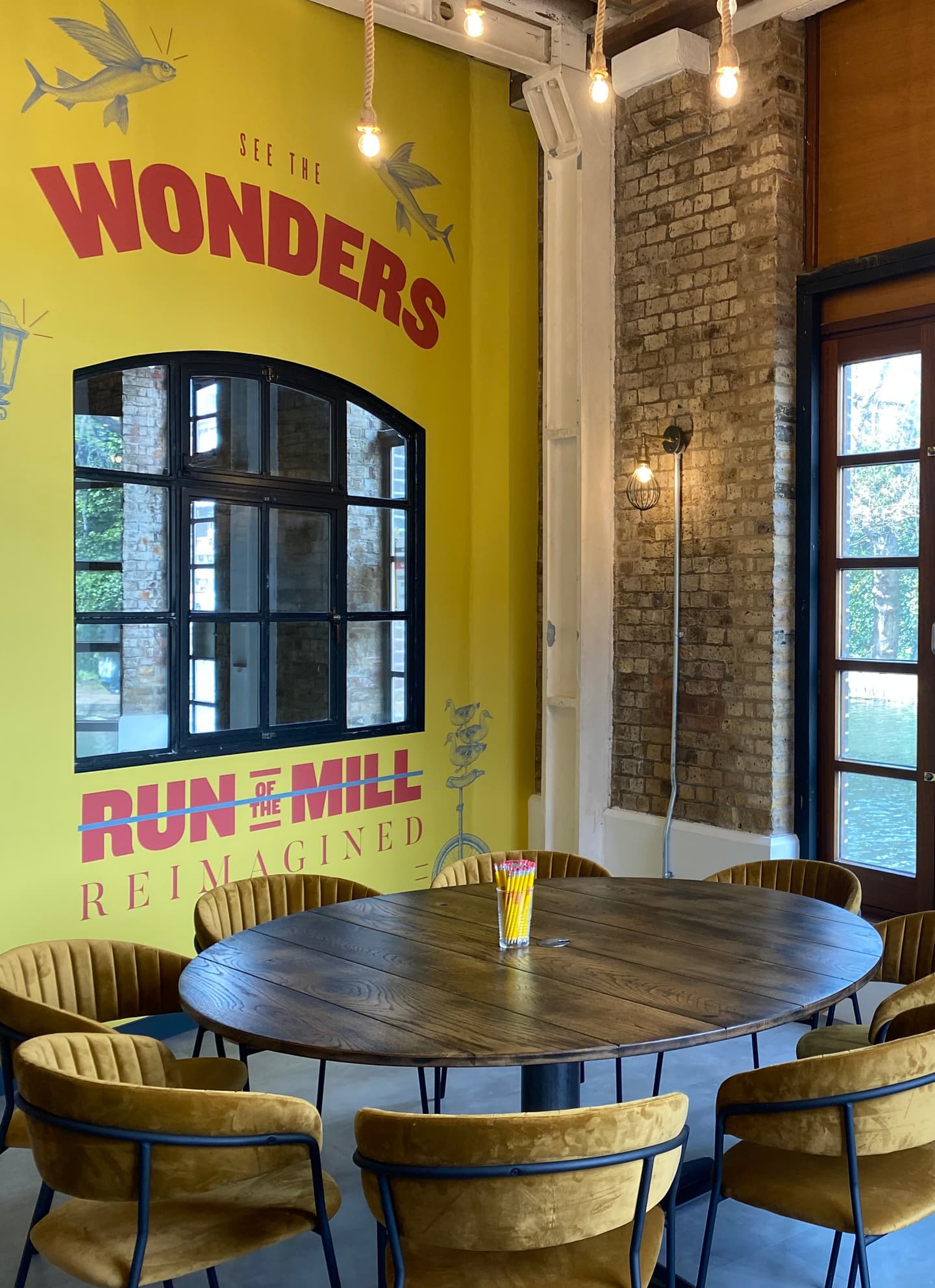 A round wooden table with six yellow chairs sits in front of a large yellow wall with bold text and a window; a jar of straws is on the table.