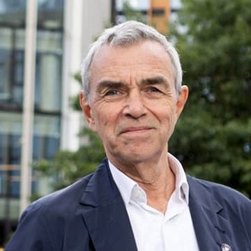 An older man with short grey hair wearing a white shirt and dark blazer stands outside in front of a modern building and greenery.