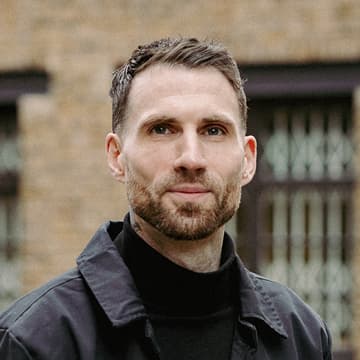 A man with short brown hair and a trimmed beard wearing a black jacket and black polo neck stands outdoors in front of a brick building with barred windows.