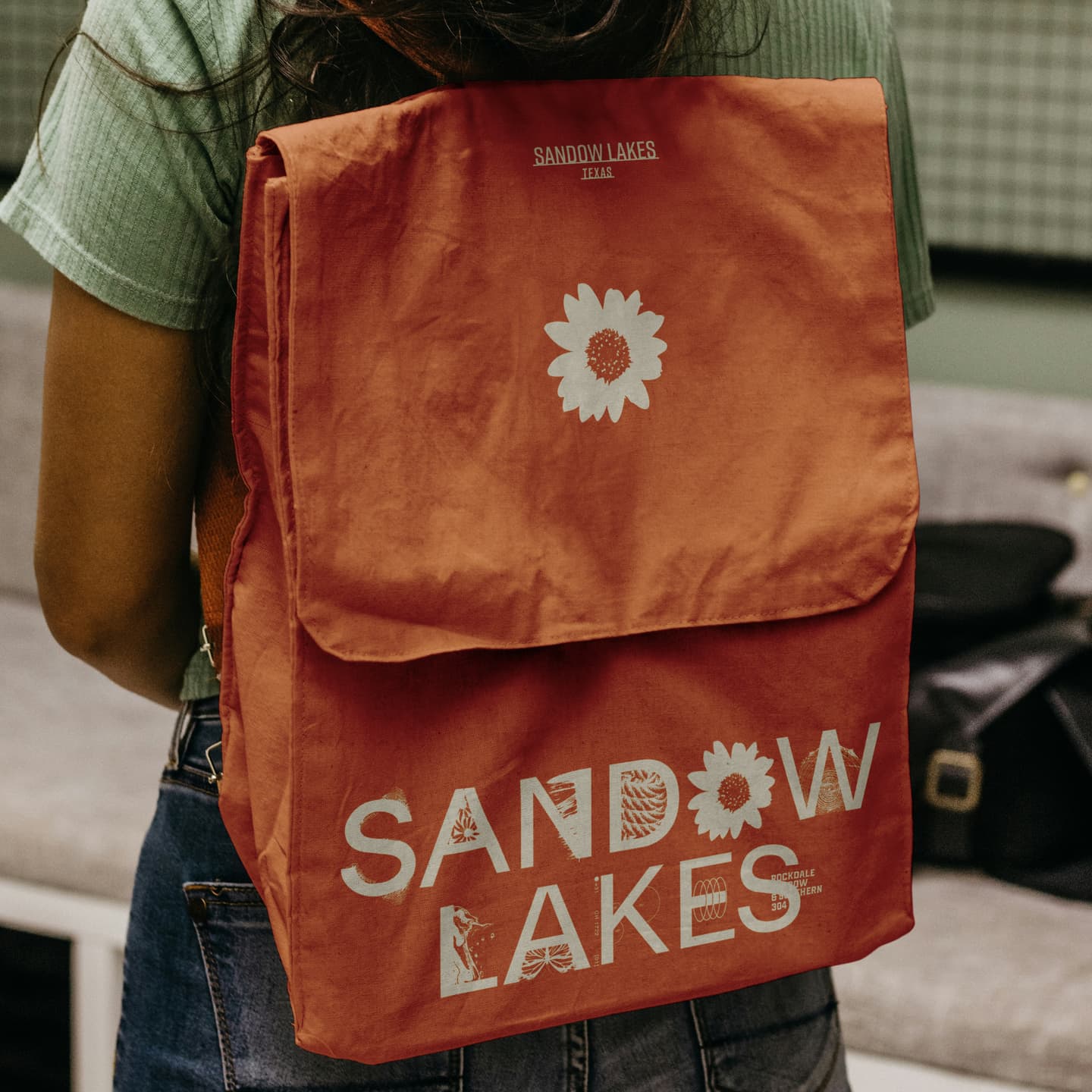 Person wearing a red rucksack with white sunflower designs and the text SANDOW LAKES printed on the flap and lower front.