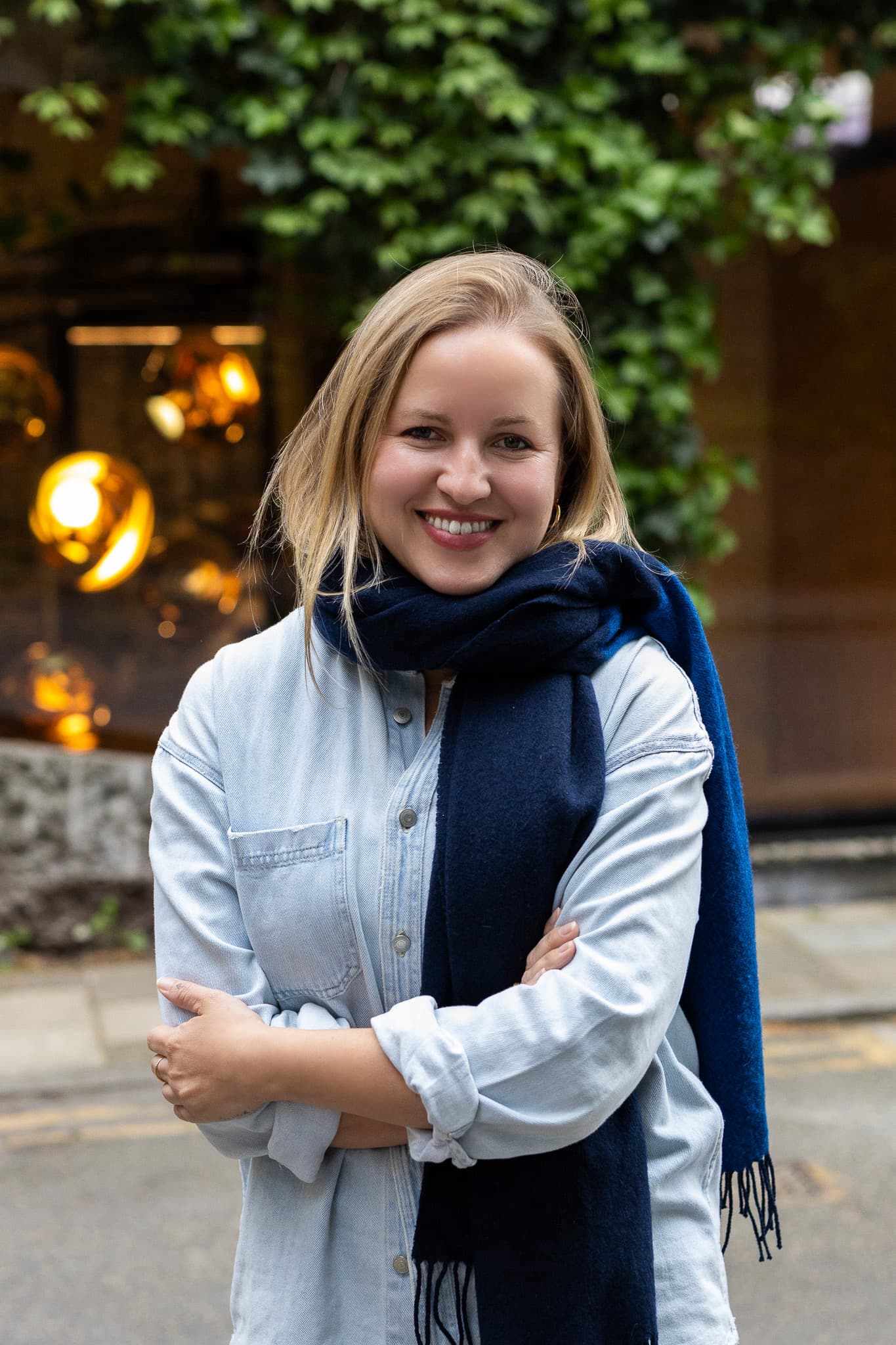 A woman with blonde hair, wearing a light denim shirt and dark blue scarf, stands outdoors smiling with arms folded. Green leaves and a window with lights are in the background.