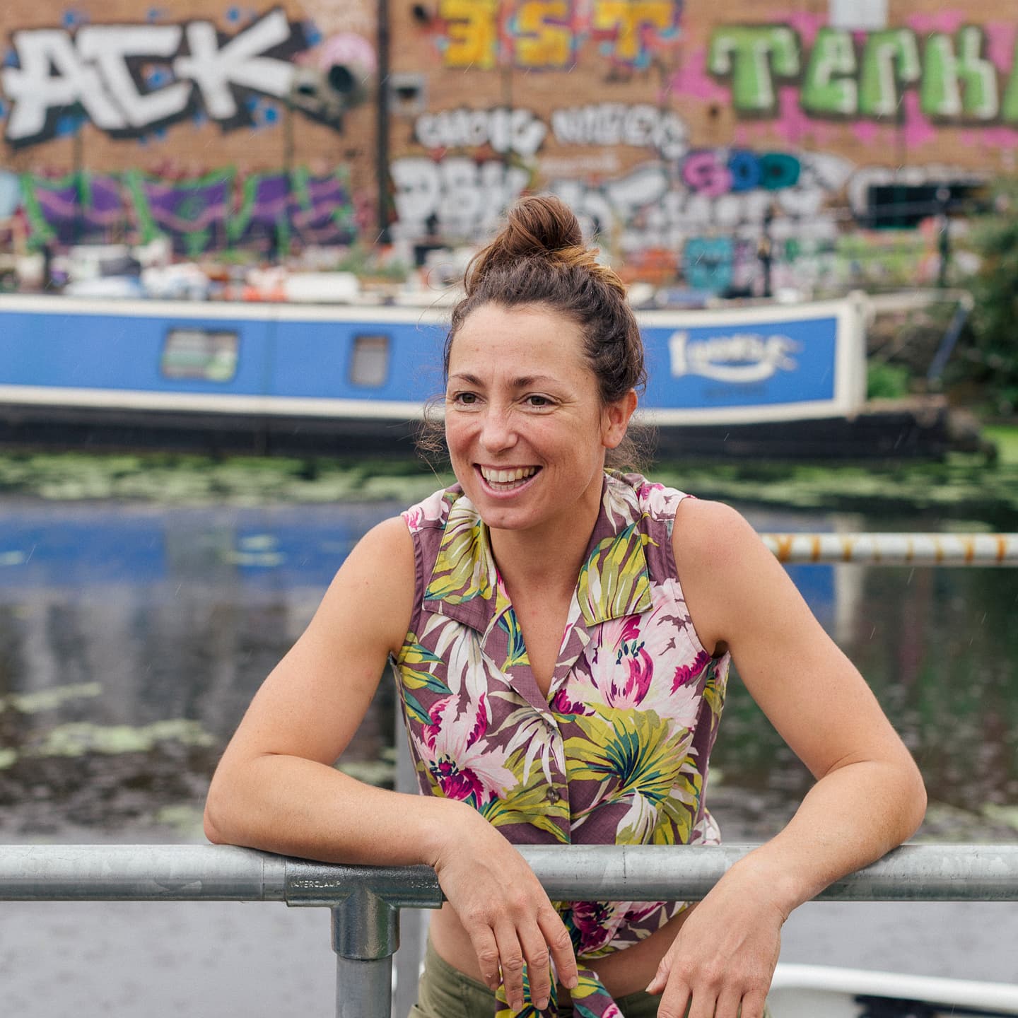 A woman in a floral shirt leans on a metal railing by a canal with a blue boat and graffiti-covered walls in the background.