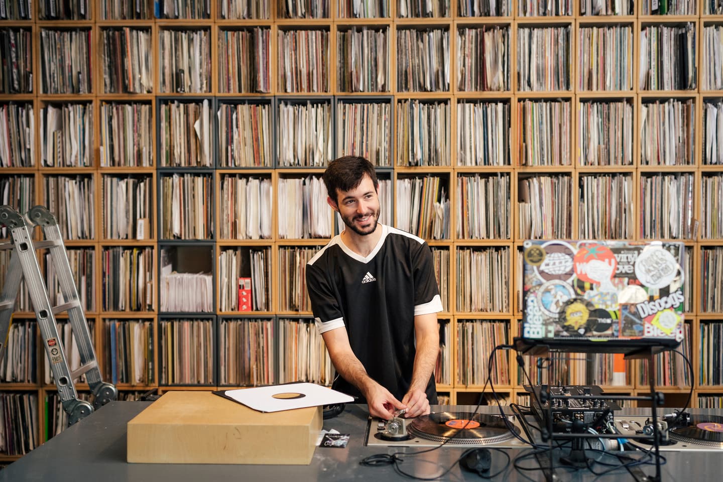 A man in a black shirt stands at a DJ set-up with vinyl records, a mixer, and a laptop, in front of shelves filled with records.