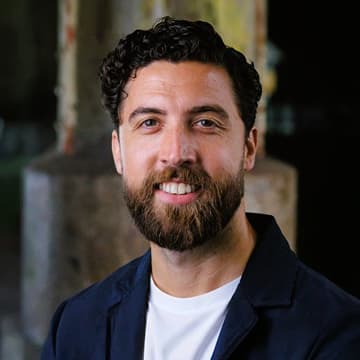 A man with dark curly hair and a beard, wearing a white shirt and dark jacket, smiles at the camera with a blurred, industrial background.