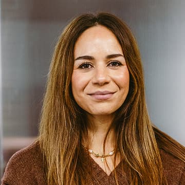 A woman with long brown hair, wearing a brown top and a necklace, smiles softly at the camera against a blurred indoor background.