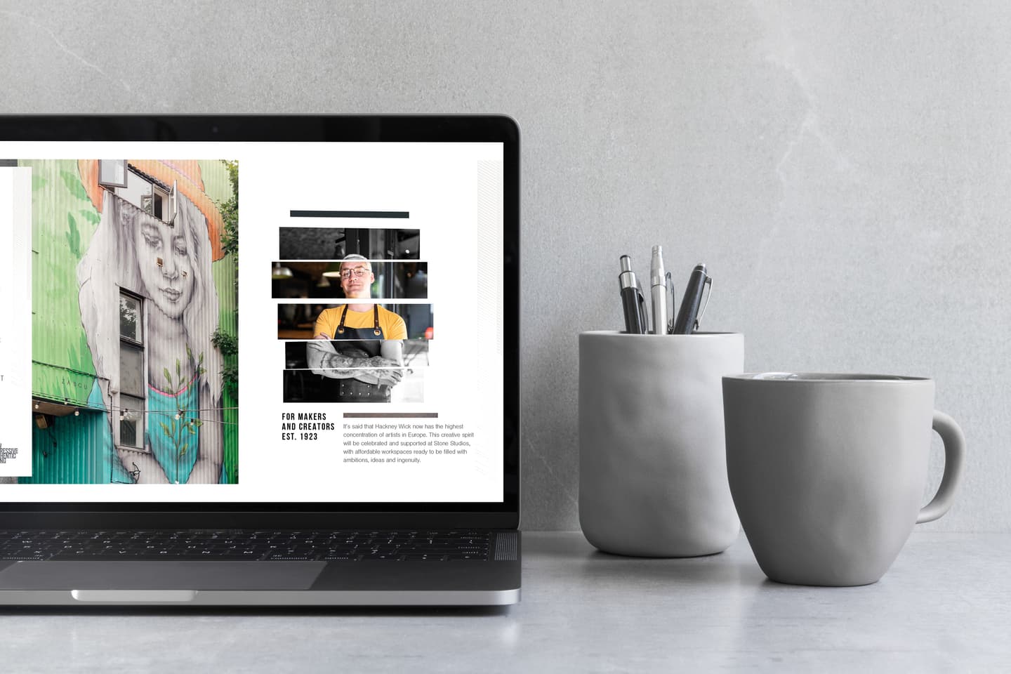 A laptop displaying an art and chef profile sits on a grey surface next to a cup holding pens and pencils and a grey mug.