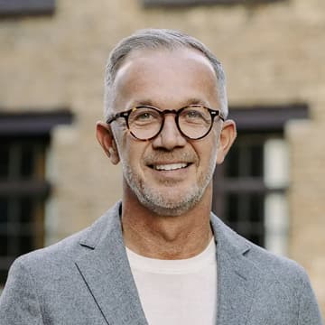 Man with short grey hair, glasses, and a beard wearing a grey blazer and white shirt, standing in front of a blurred brick building.