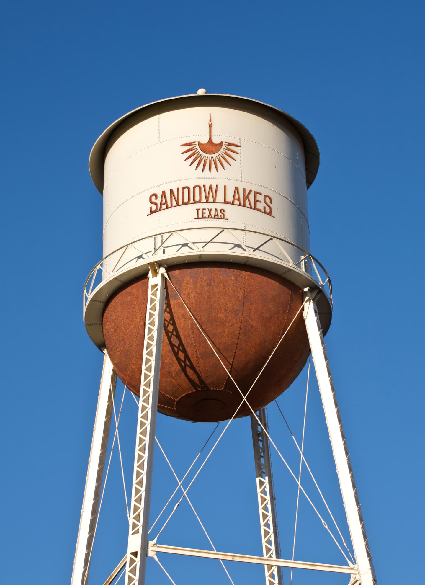 A tall white water tower with Sandow Lakes Texas painted on the tank stands against a clear blue sky.