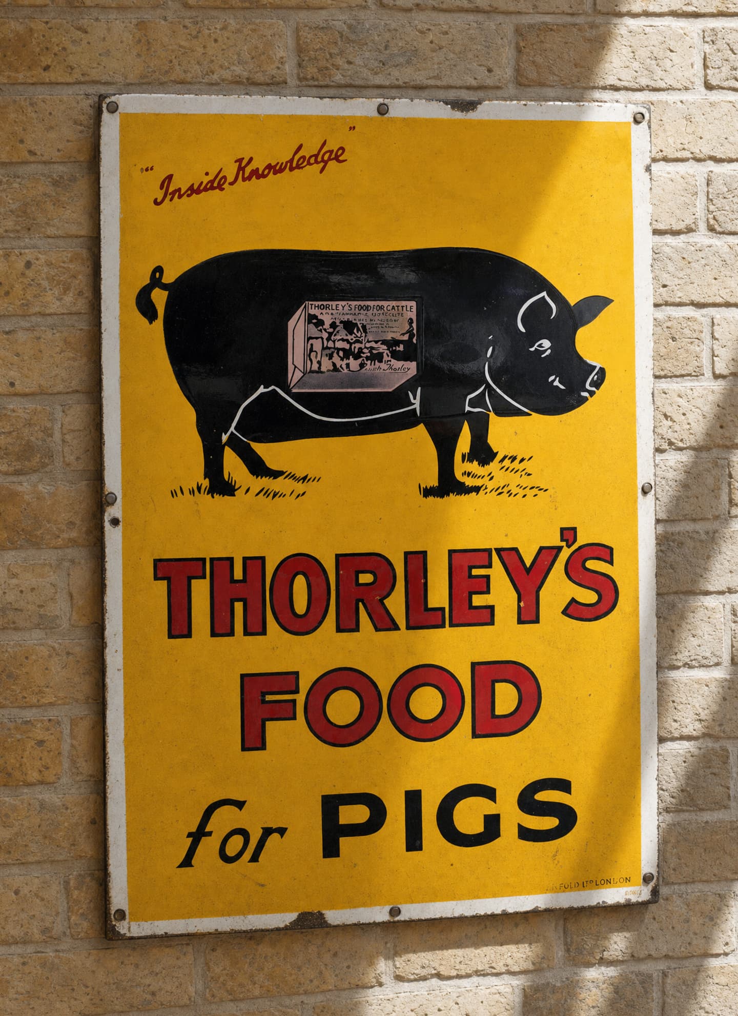 A vintage yellow metal sign on a brick wall reads Thorleys Food for Pigs with an illustration of a black pig and a feed box on its side.