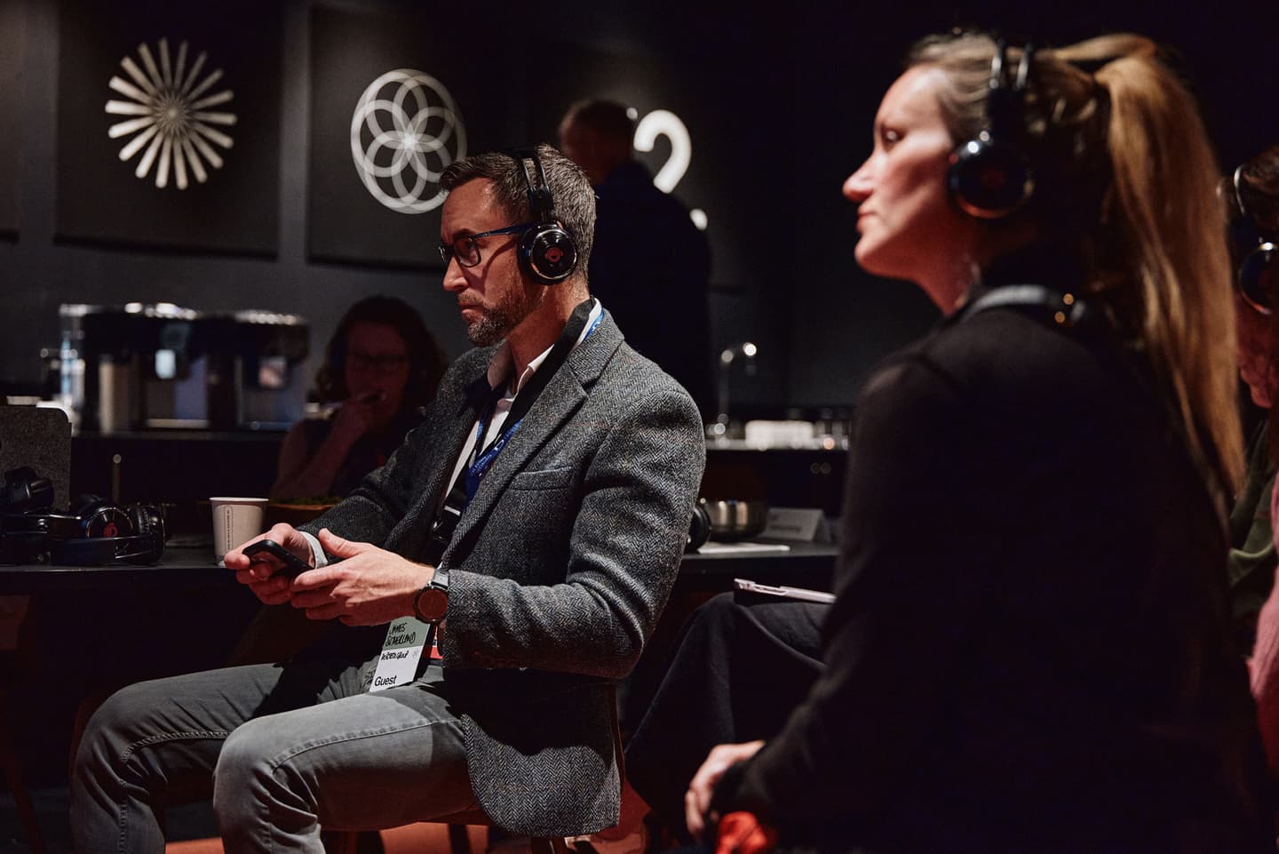 People wearing headphones sit attentively in a dimly lit room with abstract art on the walls, suggesting they are participating in an event or presentation.