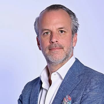 A middle-aged man with grey hair and a beard is wearing a blue suit jacket, white shirt, and pocket square, standing against a plain light background.